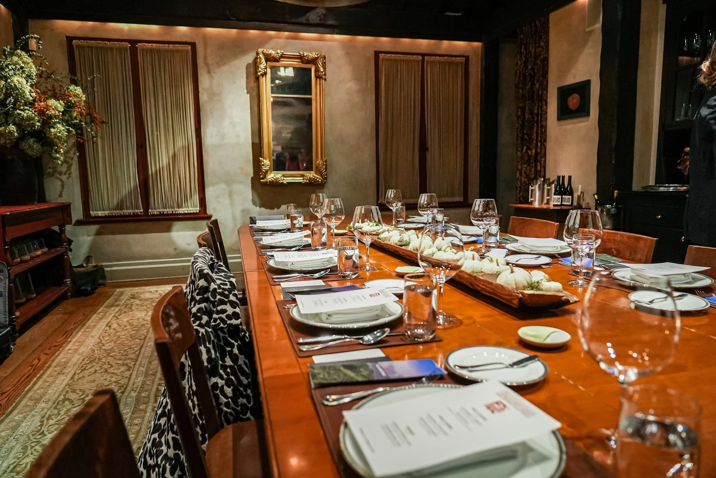A well-set dining table in a cozy restaurant with plates, wine glasses, and silverware, decorated with a long tray of pumpkins and gourds for fall or Thanksgiving, amid warm lighting and elegant decor.