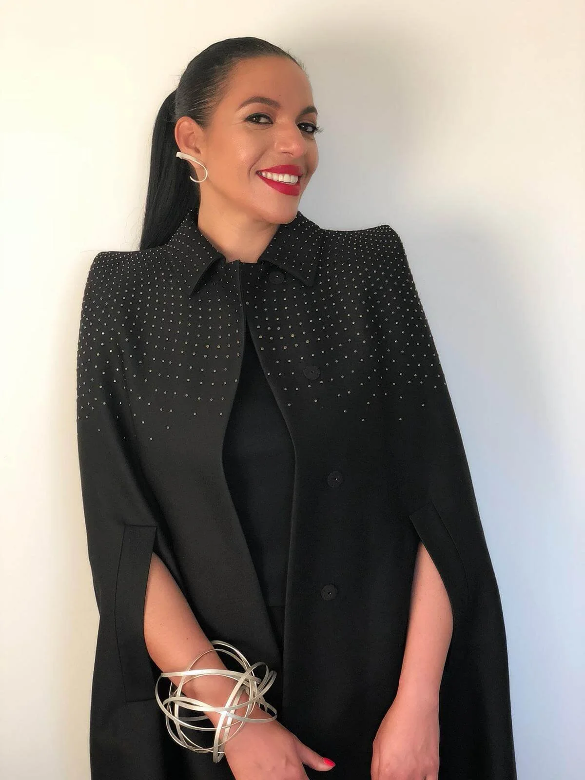 A woman with long dark hair wearing a black dress and a black jacket with white polka dots, smiling against a plain white background.