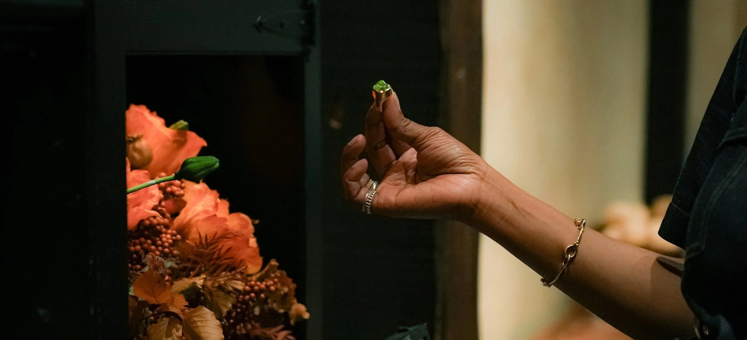 A person's hand holding a small green item, with floral arrangements in the background.