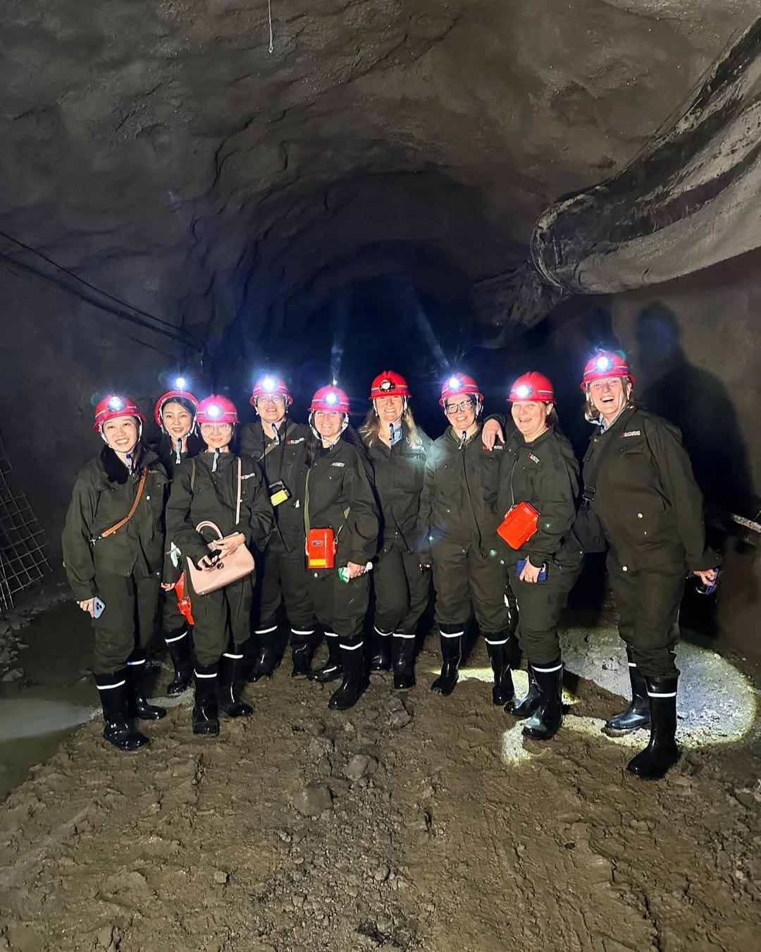 A group of nine women dressed in caving gear, including helmets with headlamps, standing inside a cave with rugged walls and dirt floor, smiling for a photo.