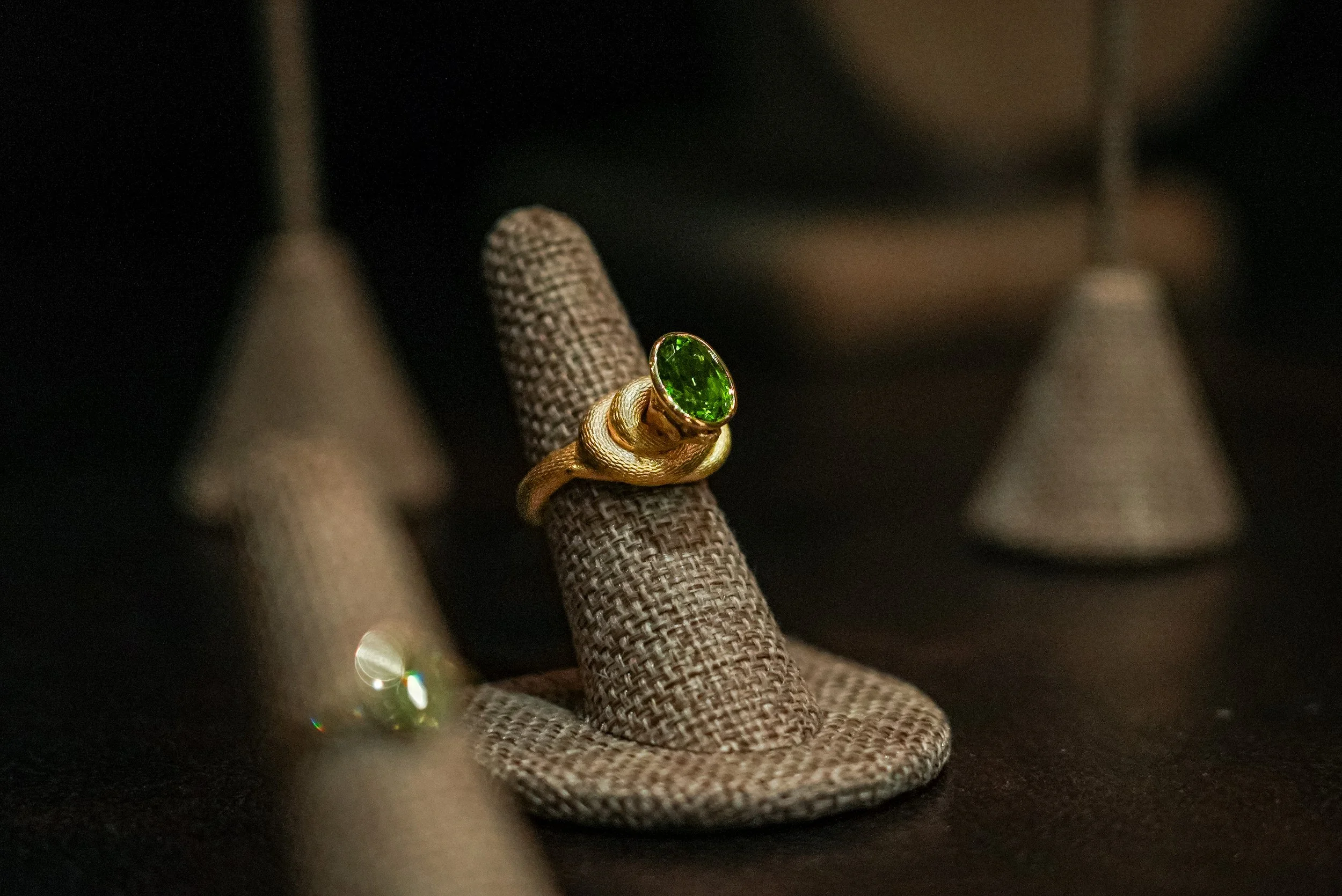 Gold ring with a large green gemstone displayed on a fabric ring holder, surrounded by other jewelry displays in a dark background.
