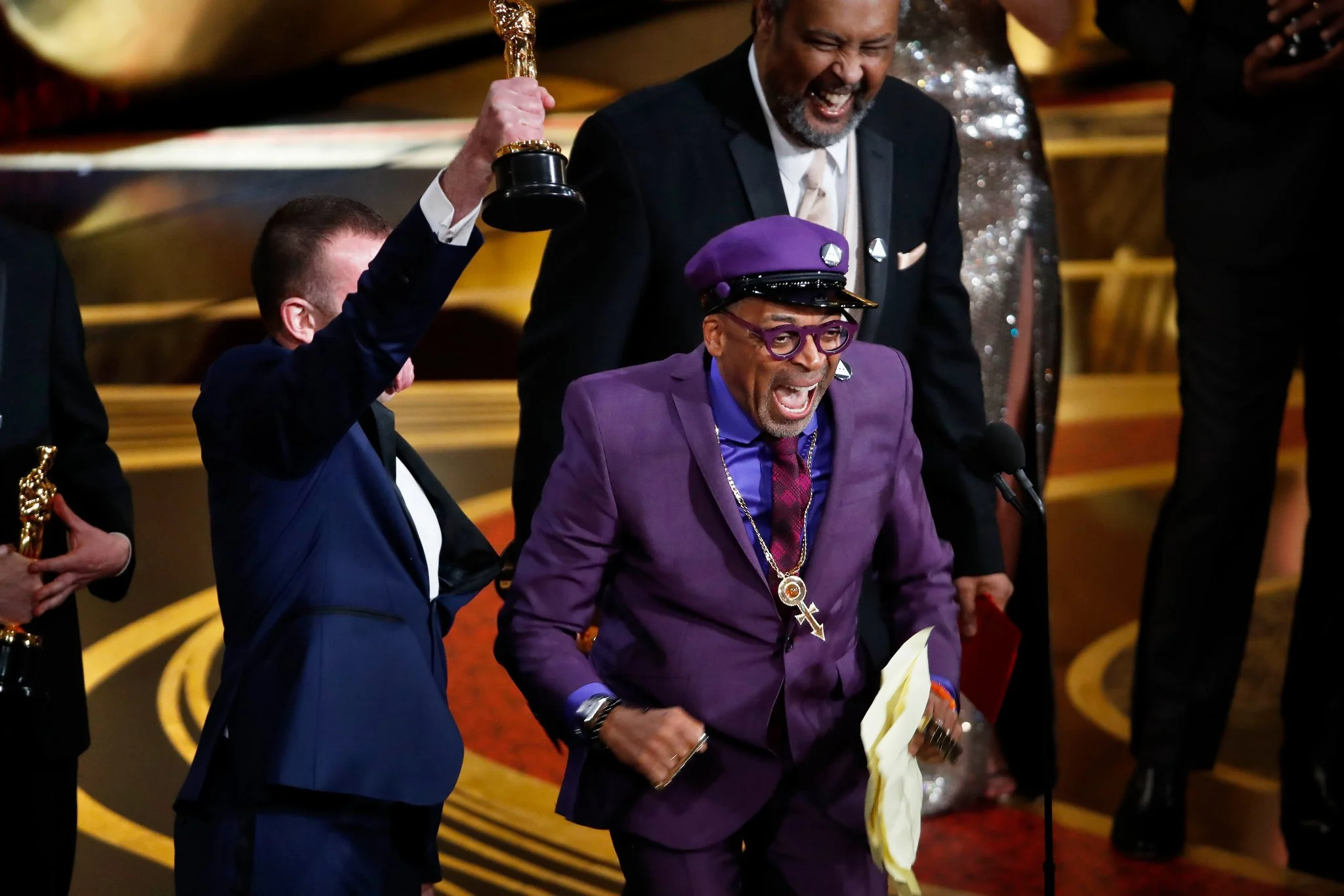 An award ceremony with three men celebrating on stage, one in a purple suit and hat, holding a yellow cloth, and wearing glasses, jewelry, and a necklace with a key and cross. The man in the purple suit is smiling and appears excited, while a man in a navy suit, next to him, raises an award in his hand. The third man behind them is dressed in a black suit and laughing.