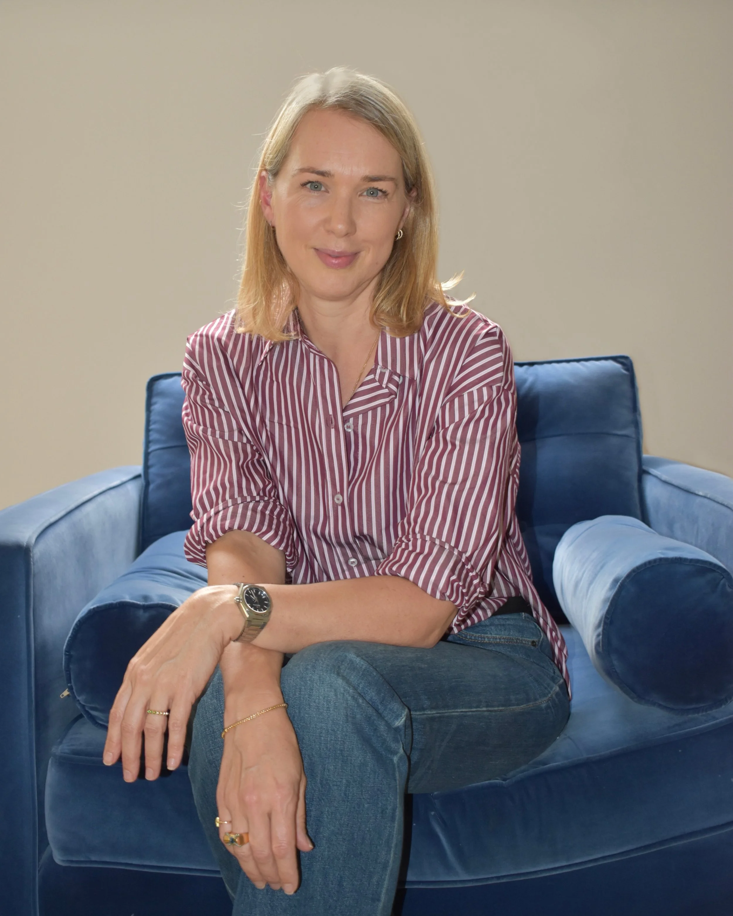 A woman with blonde hair, wearing a striped shirt, sitting on a blue velvet armchair with a neutral background.