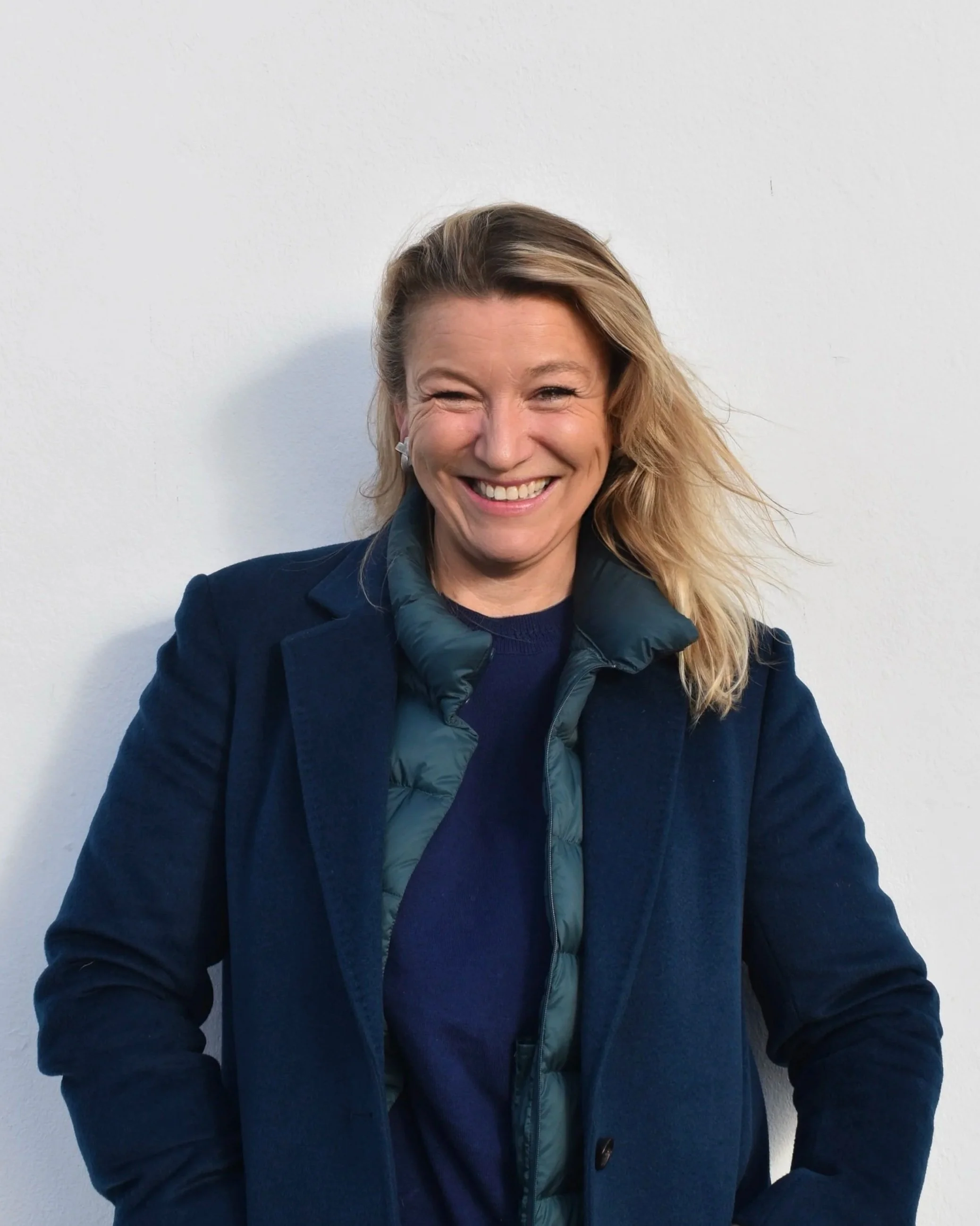 A woman smiling, wearing a navy blue blazer and a dark green puffer vest, standing against a white wall.