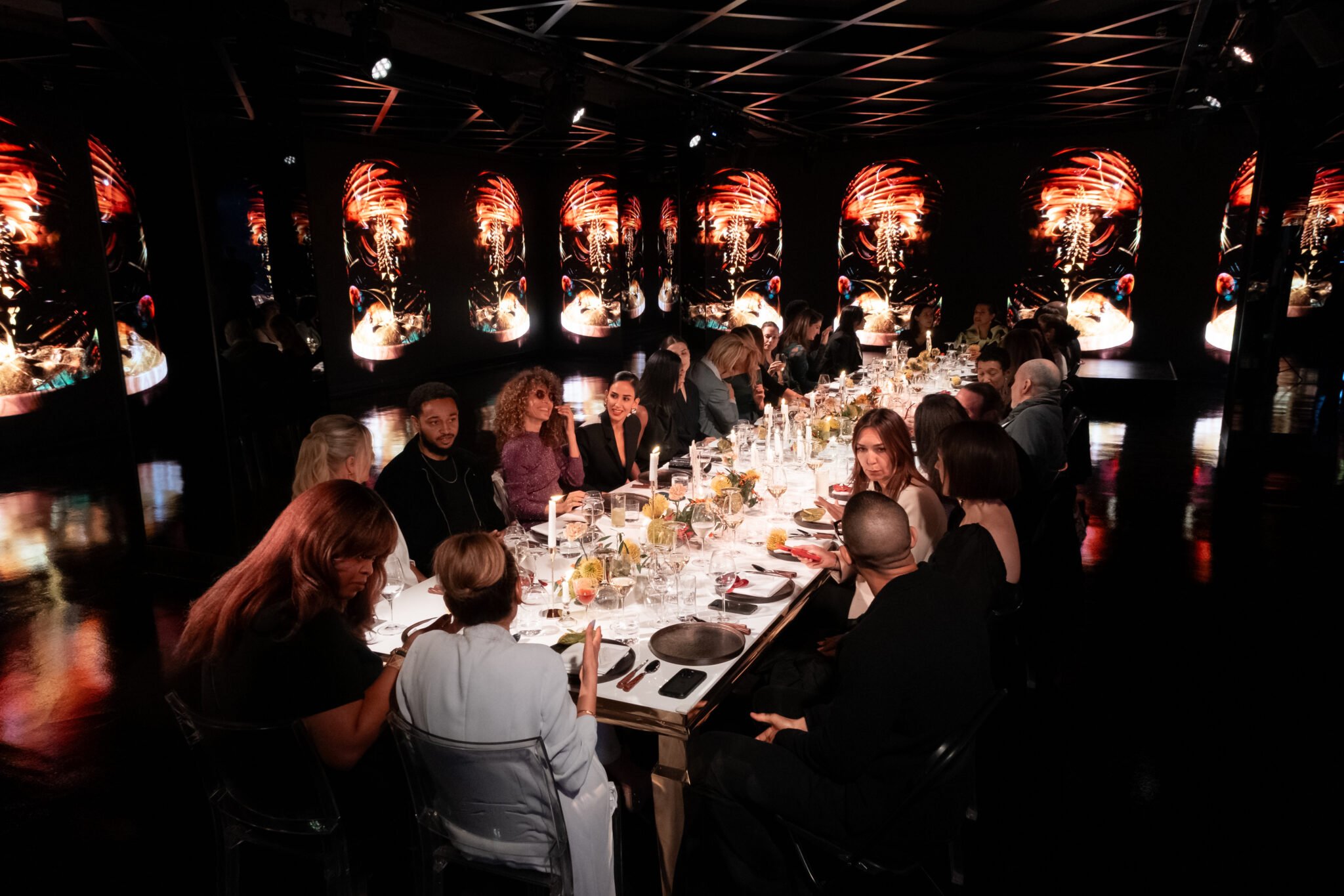People gathered around a long dinner table in a dark room with colorful abstract digital artwork on the walls.