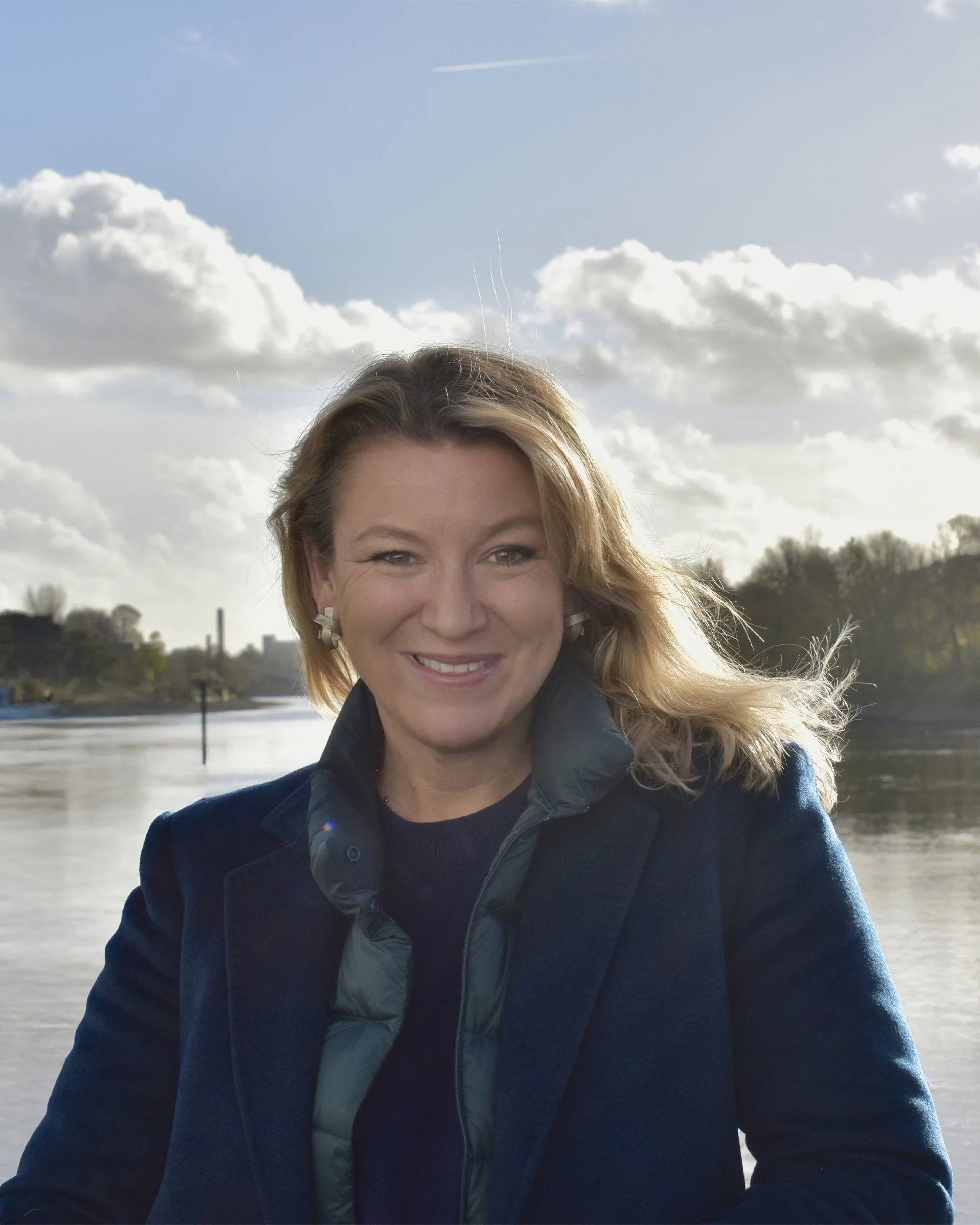 A woman outdoors smiling with a river and cloudy sky in the background.