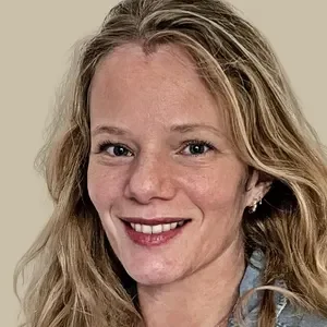 Close-up portrait of a woman with long wavy blond hair smiling at the camera against a plain background.