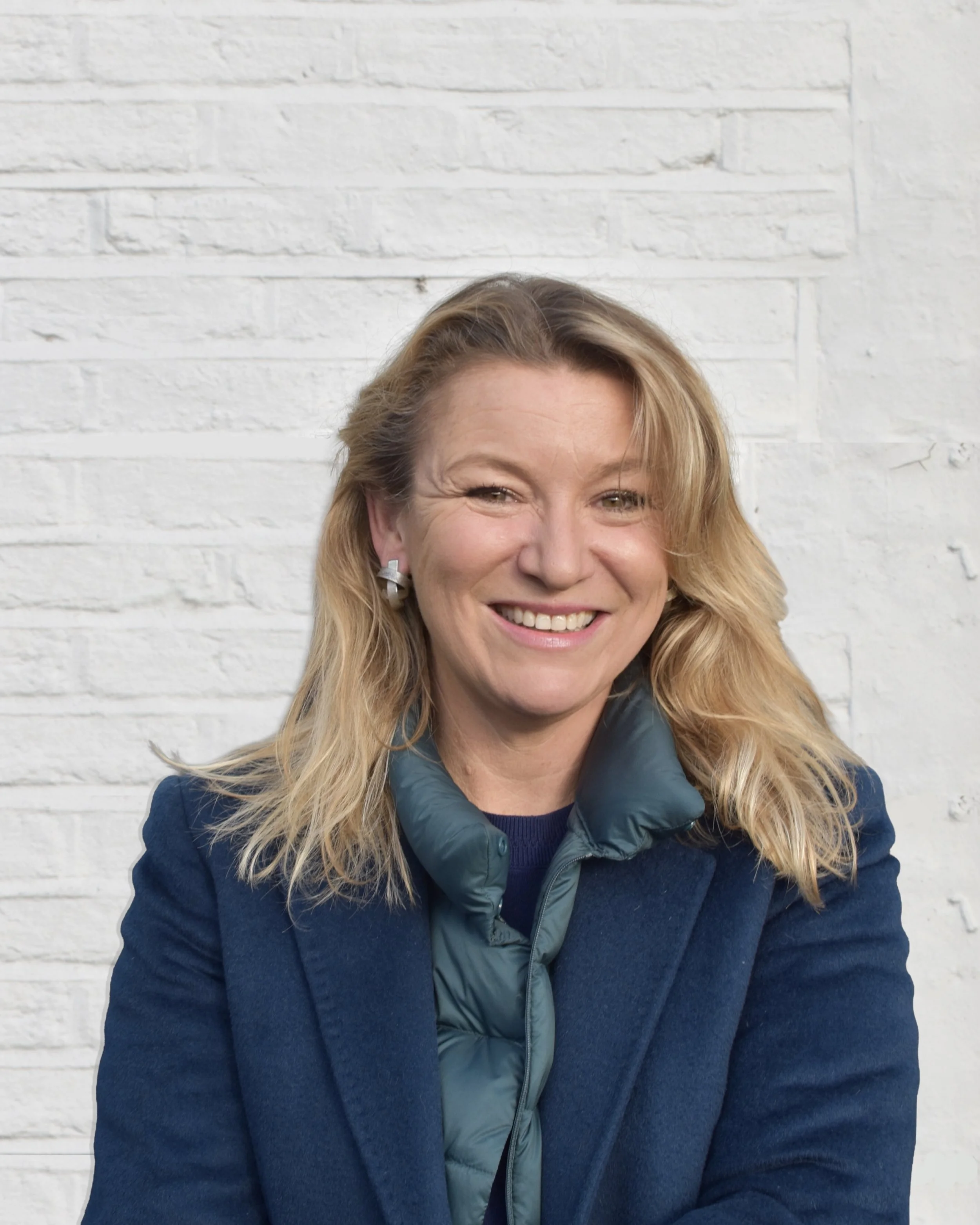 A smiling woman with shoulder-length blonde hair, wearing a dark blue coat and a green puffer jacket, standing against a white brick wall.