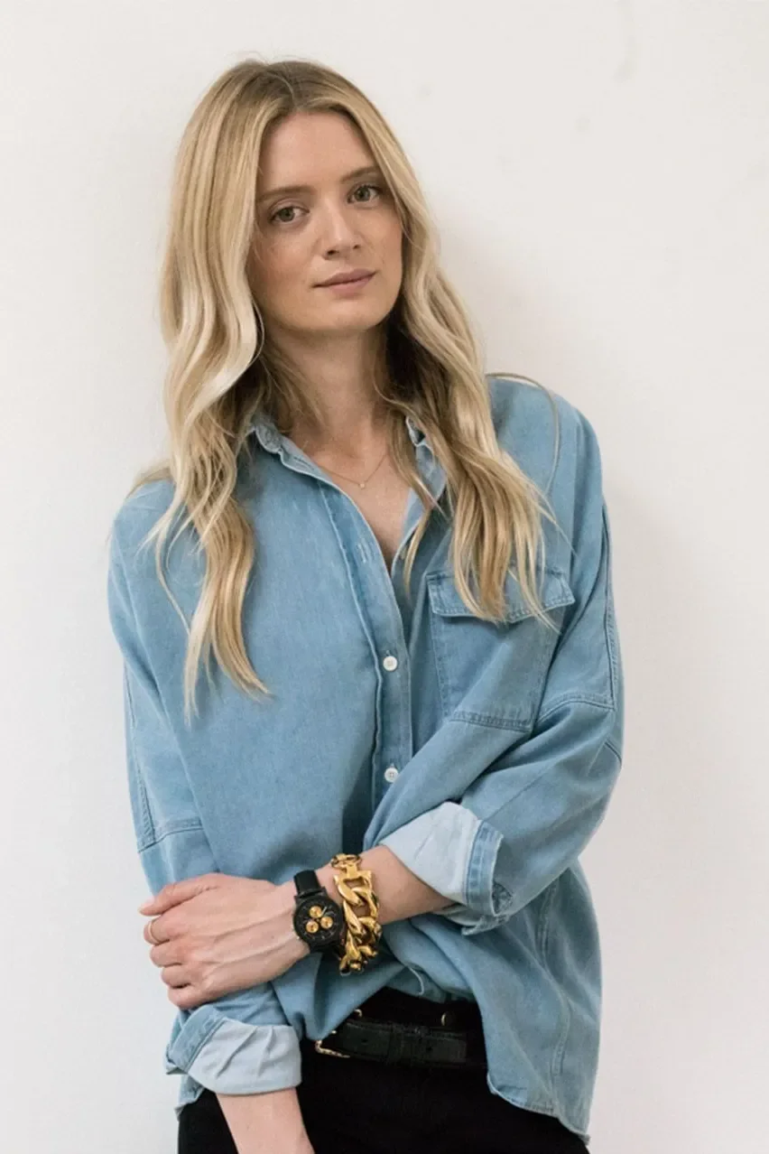 A woman with long blonde hair wearing a loose-fitting denim shirt and black pants, standing against a white wall.