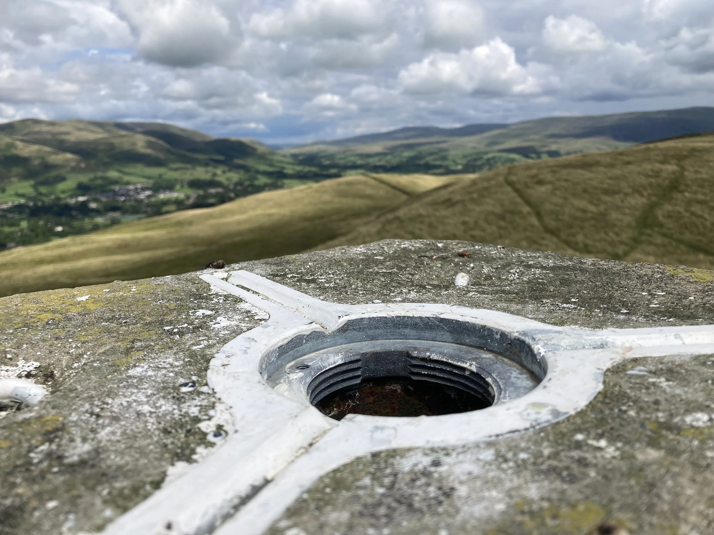 Close-up of a weathered stone summit marker with an embedded metal screw, overlooking rolling green hills and a valley under a partly cloudy sky.