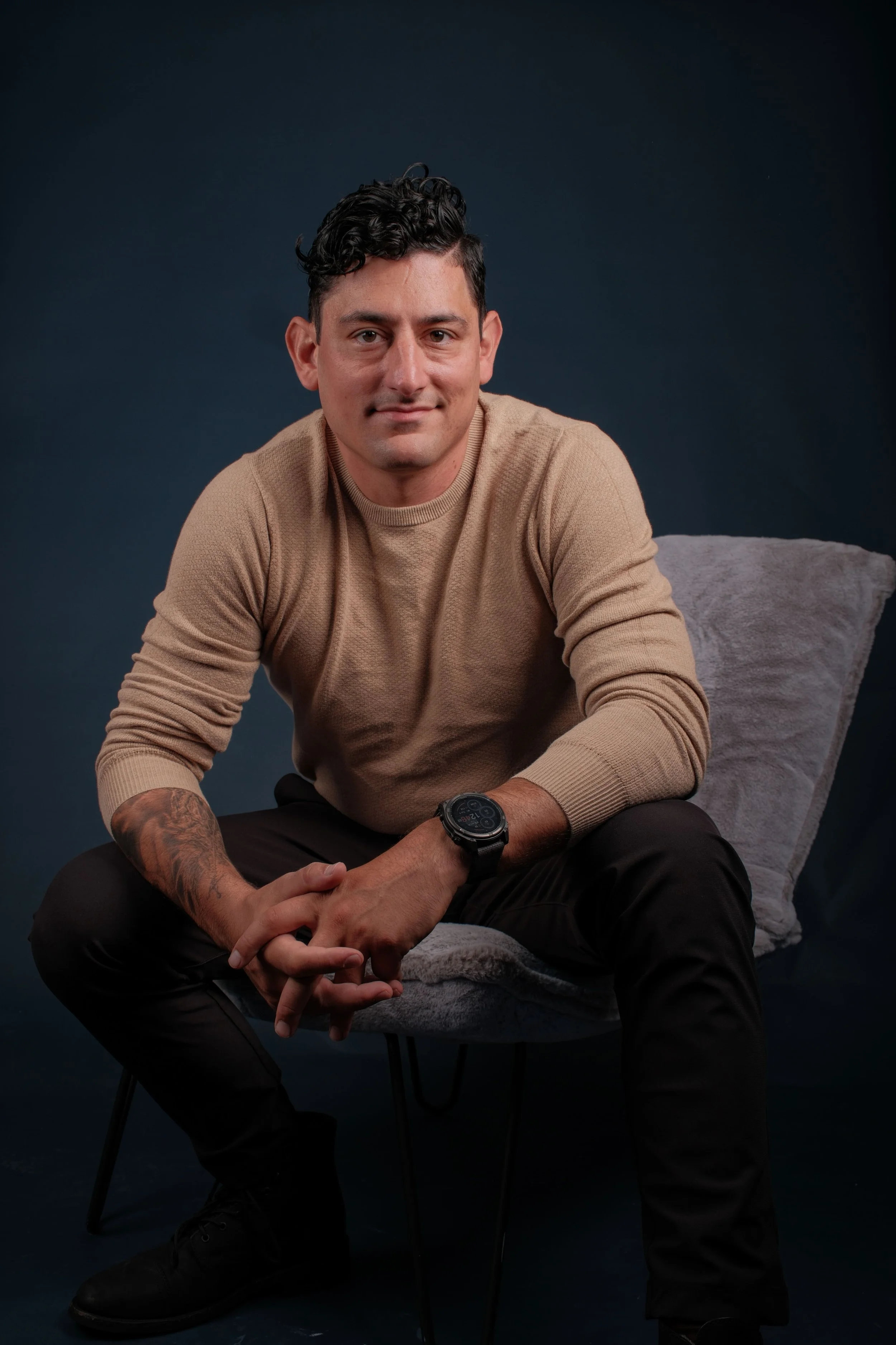 Alex Rodino, founder of Rodino Creative Works, seated in a beige sweater and dark pants — representing mindful, purpose-driven leadership built through experience and clarity.