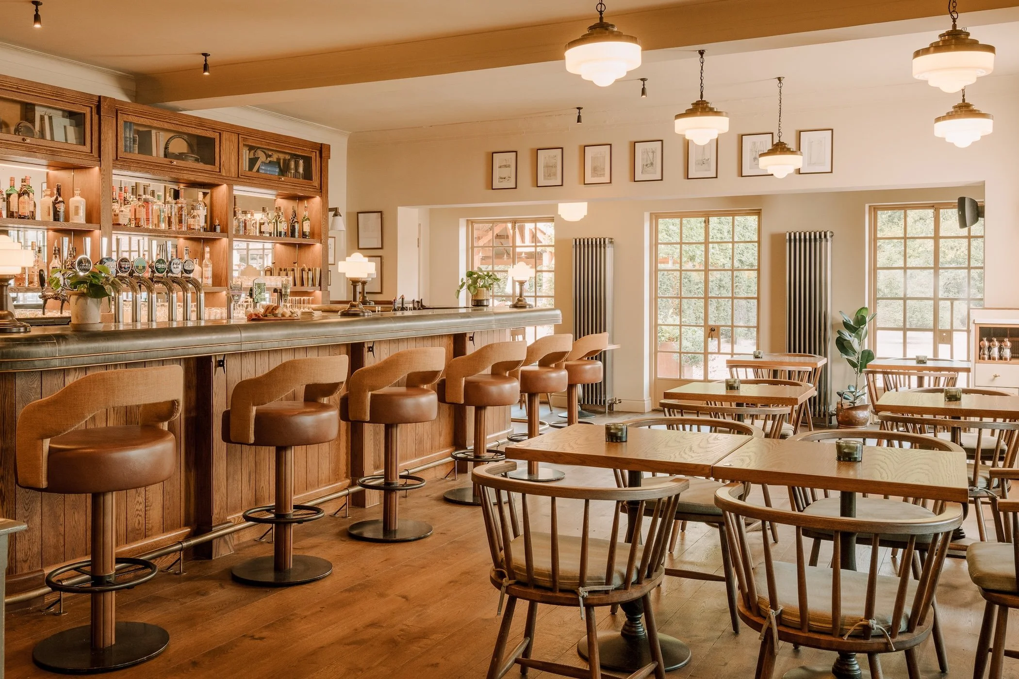 Empty restaurant with a wooden bar, barstools, and tables, illuminated by hanging pendant lights and lamps, featuring large windows, framed art on the walls, and potted plants.
