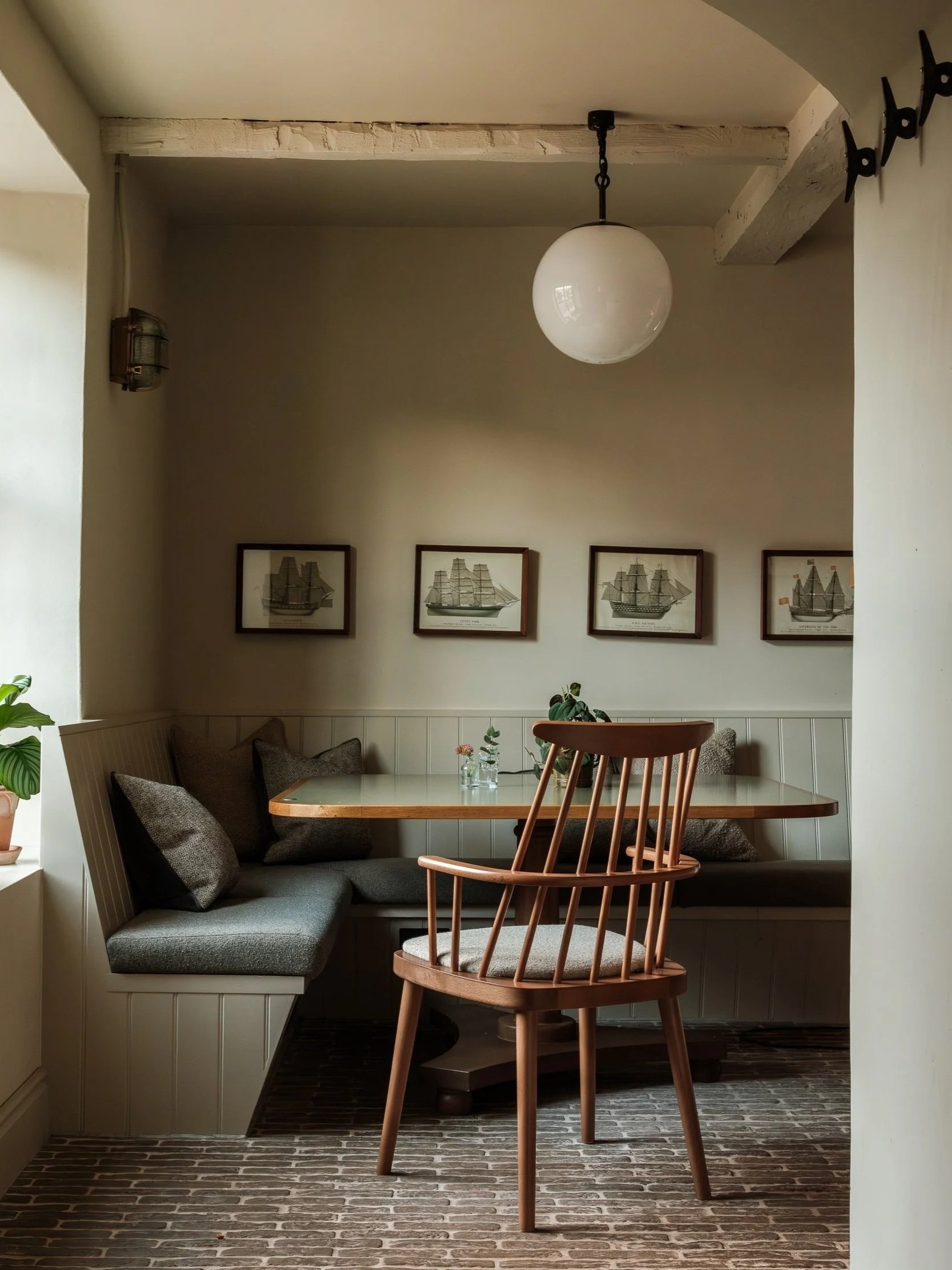 Cozy dining nook with a wooden window seat, dark gray cushions, and throw pillows, a round wooden table, a modern wooden chair, framed ship sketches on the wall, a globe-shaped pendant light hanging from the ceiling, potted plants on the window sill, and a brick-patterned floor.