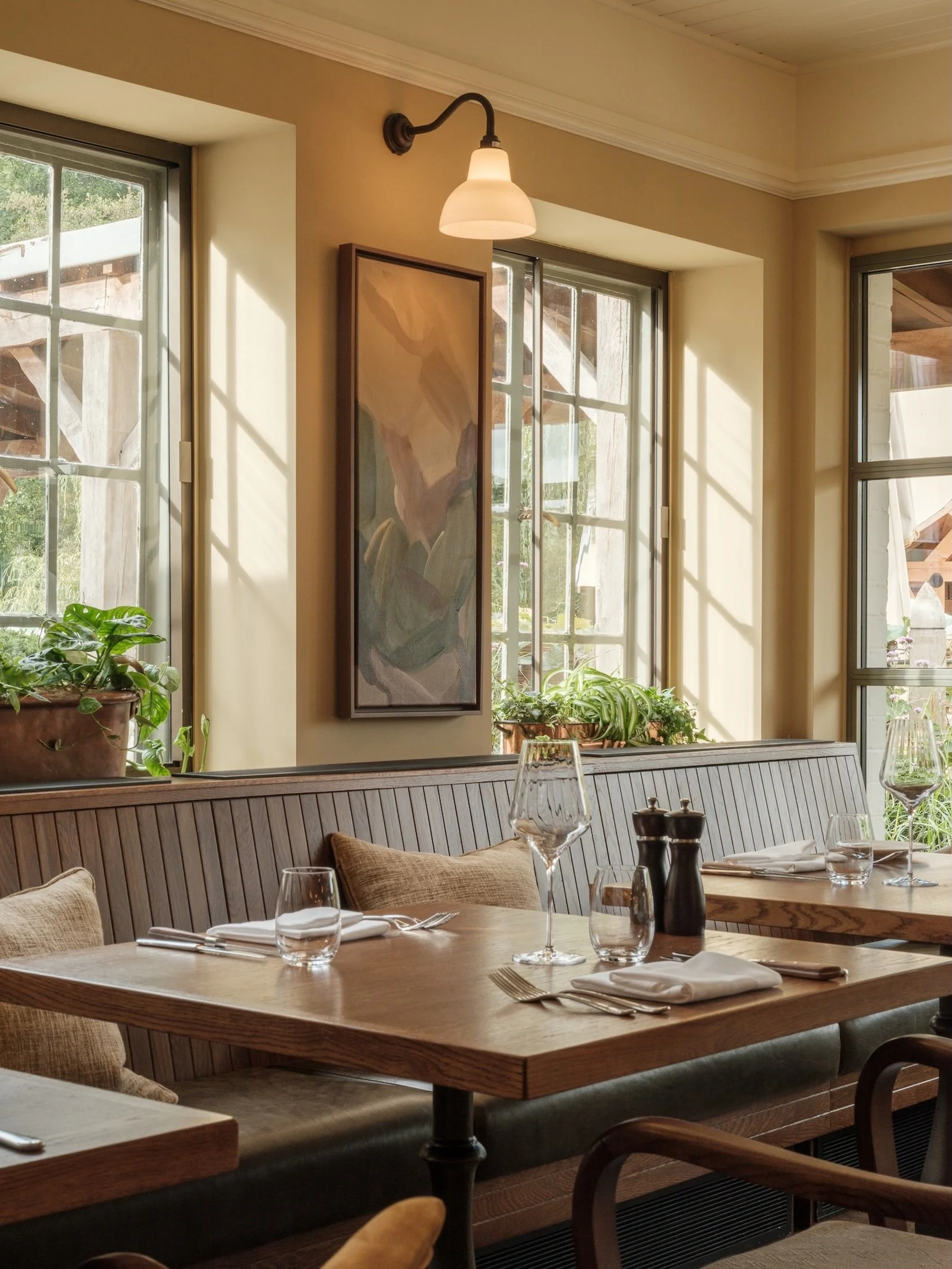 A cozy restaurant corner with wooden tables set with wine glasses, water glasses, napkins, and cutlery, a wall-mounted light, and potted plants beside large windows with sunlight streaming in.