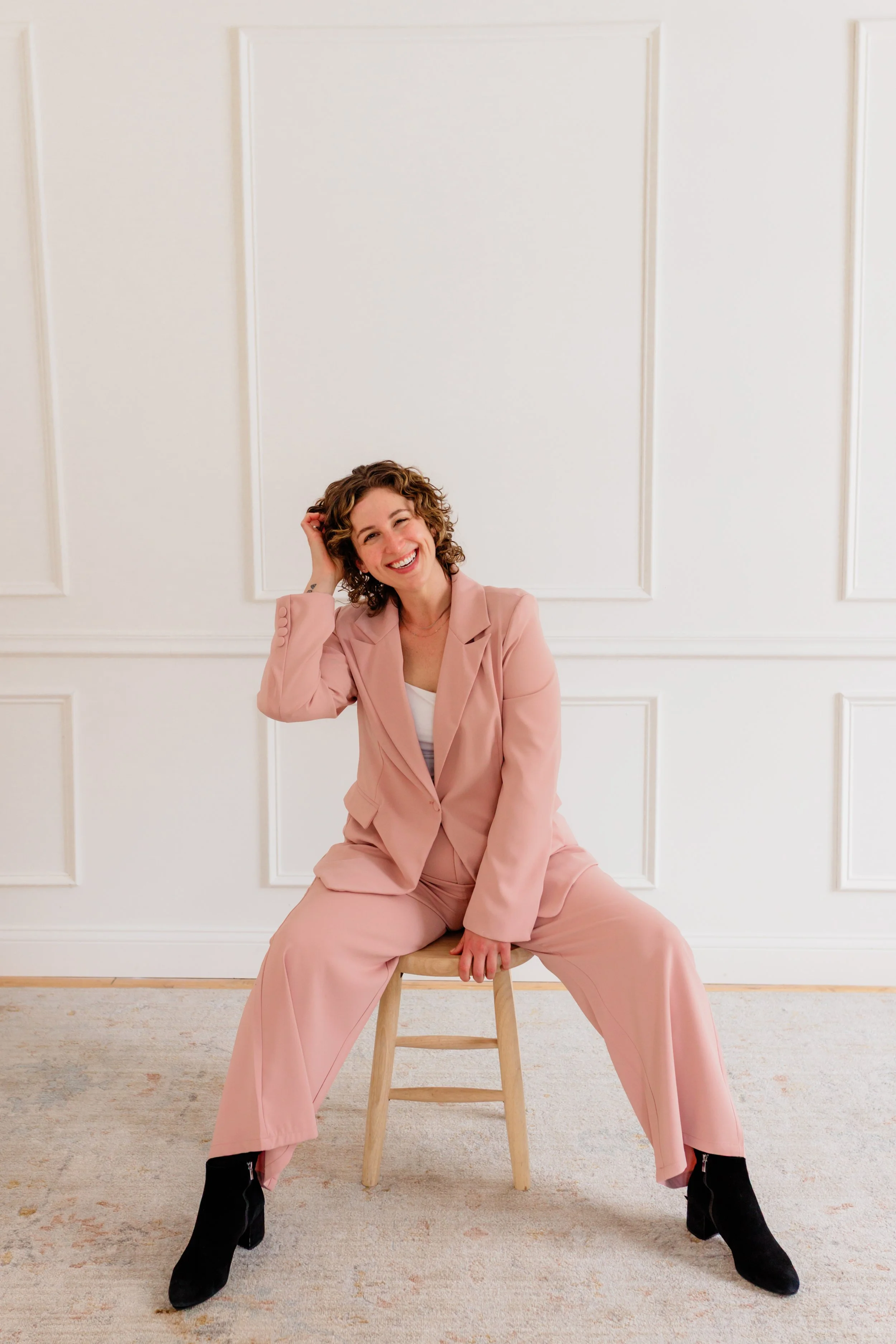 Woman with curly hair sitting on a wooden stool, smiling, wearing a pink suit with black ankle boots, in front of a white paneled wall.
