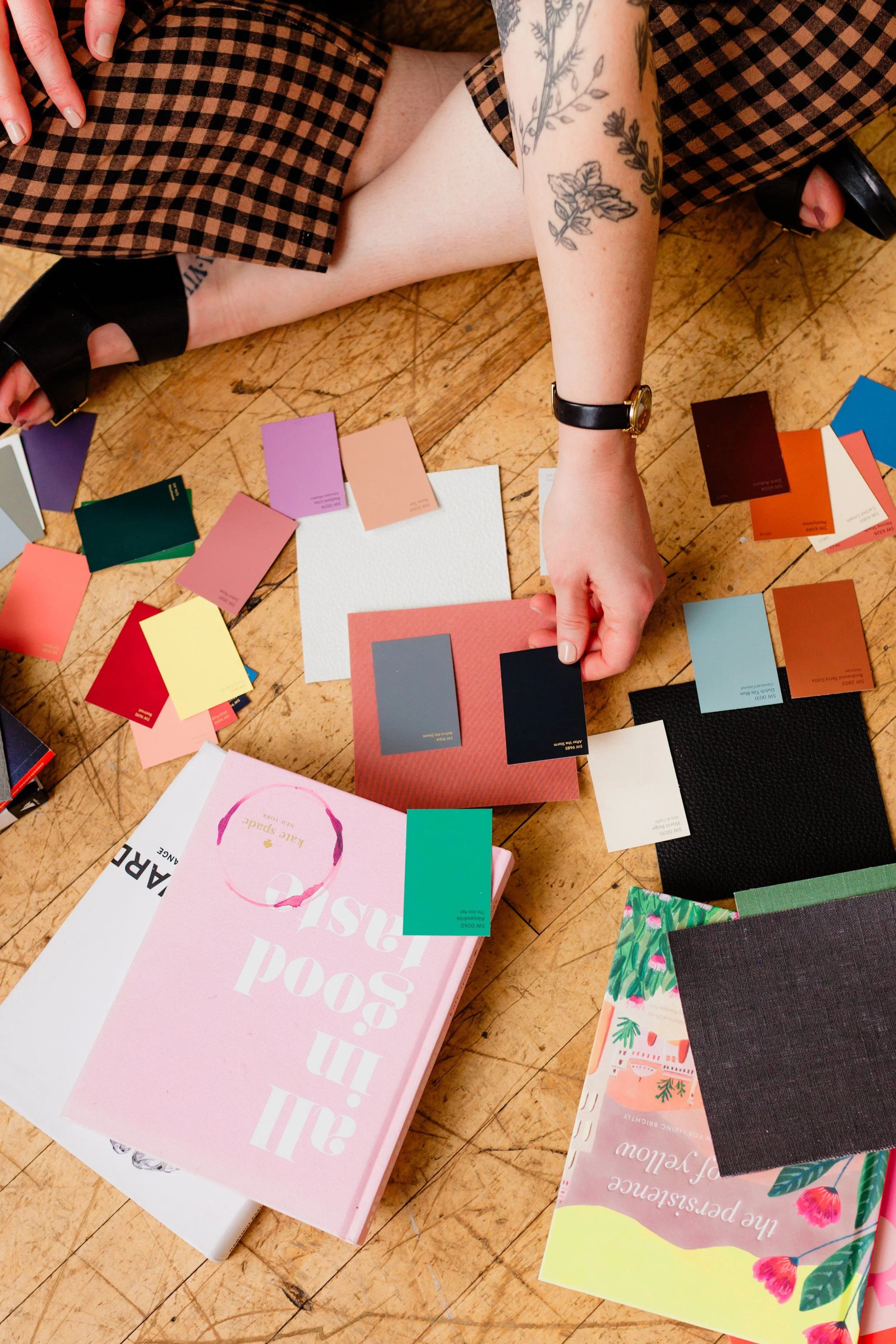 Person sitting on wooden floor, touching color swatch cards and sample books, with painted arm tattoos, wearing a checkered dress and black sandals.