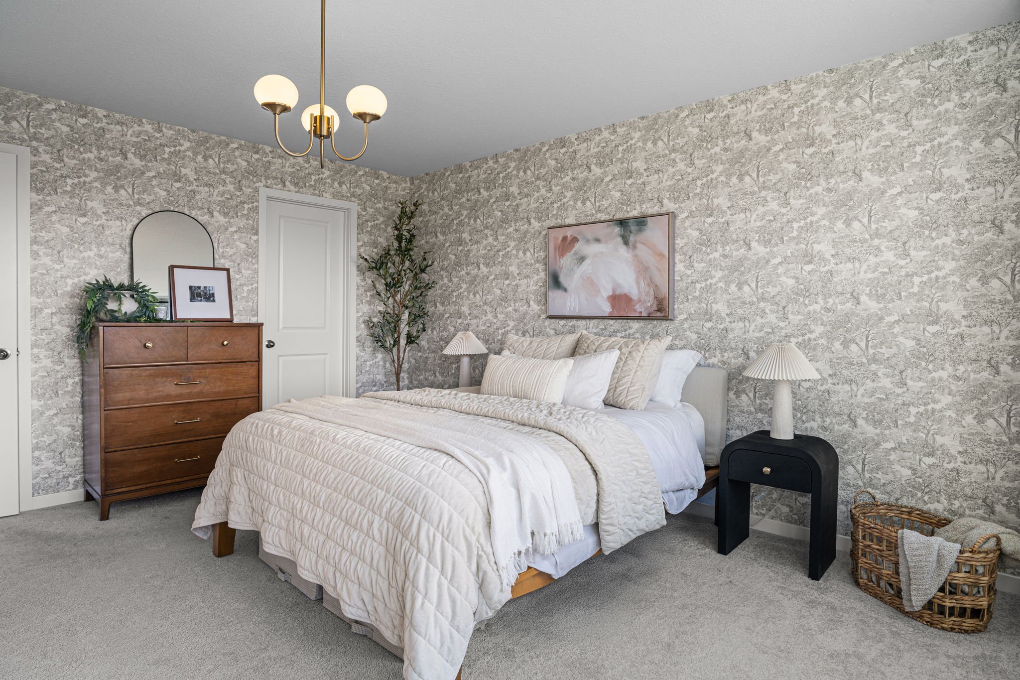 A neatly made bed with a cream-colored quilt, multiple pillows, and a headboard against a wallpapered wall with a floral pattern. There are two bedside tables with lamps and a framed artwork hanging above the bed. A wooden dresser with a mirror, fram