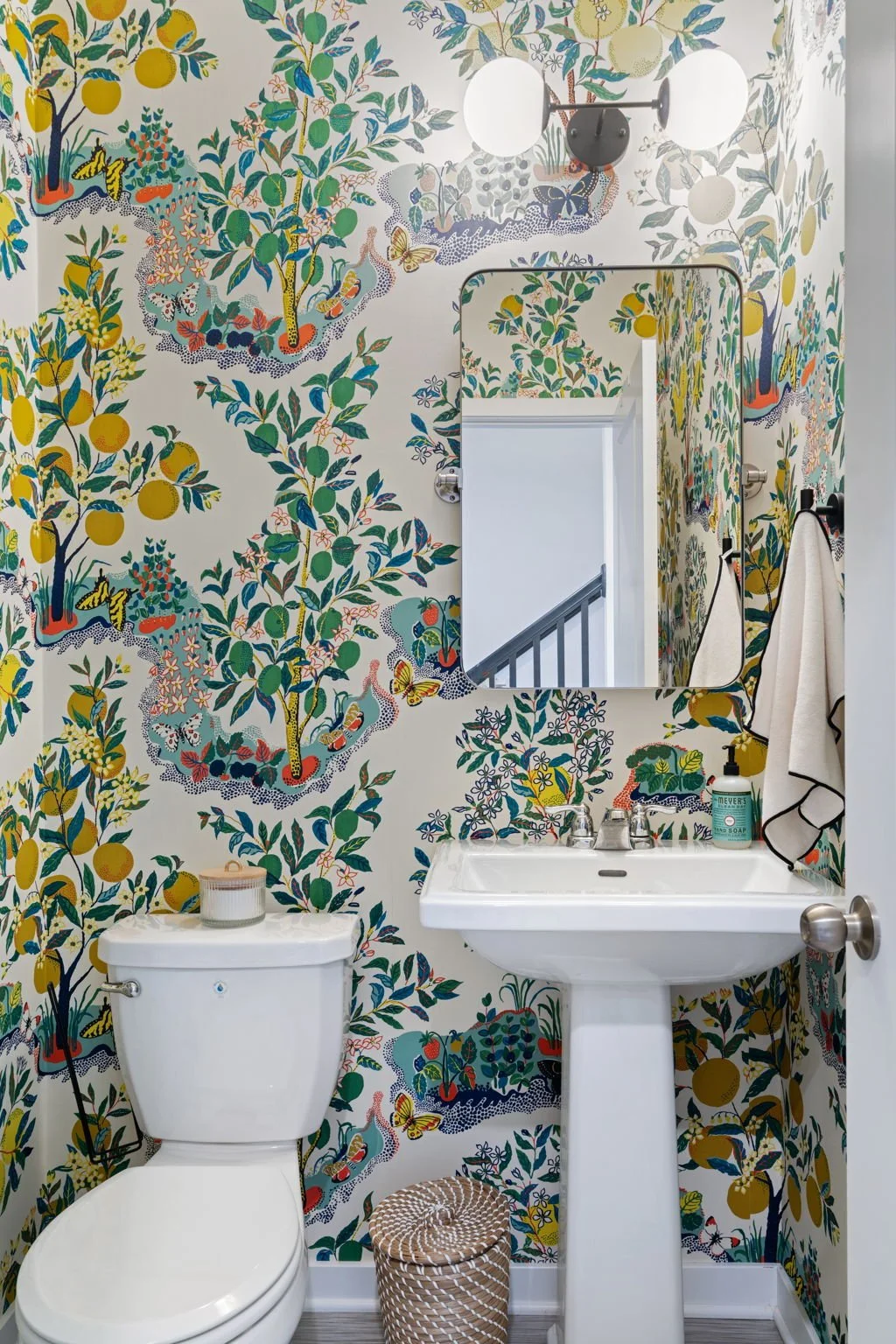 A small bathroom with vibrant, patterned wallpaper featuring lemon trees, butterflies, and flowering plants. There is a white toilet and a white pedestal sink with a mirror above it, reflecting a stairway. A soap dispenser and hand towel are visible 