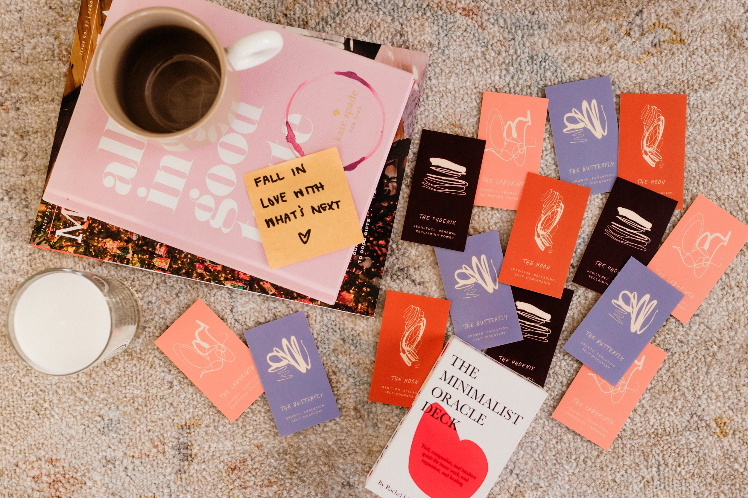 A collection of tarot or oracle cards spread out on a beige carpet, with a book titled 'The Minimalist Oracle Deck', a pink notebook with a sticky note saying 'Fall in love with what’s next', a coffee mug, and a glass of milk. The cards are in various colors including black, purple, orange, and pink, each with abstract line art and titles such as 'The Moon', 'The Butterfly', 'The Phoenix', and 'The Labyrinth'.