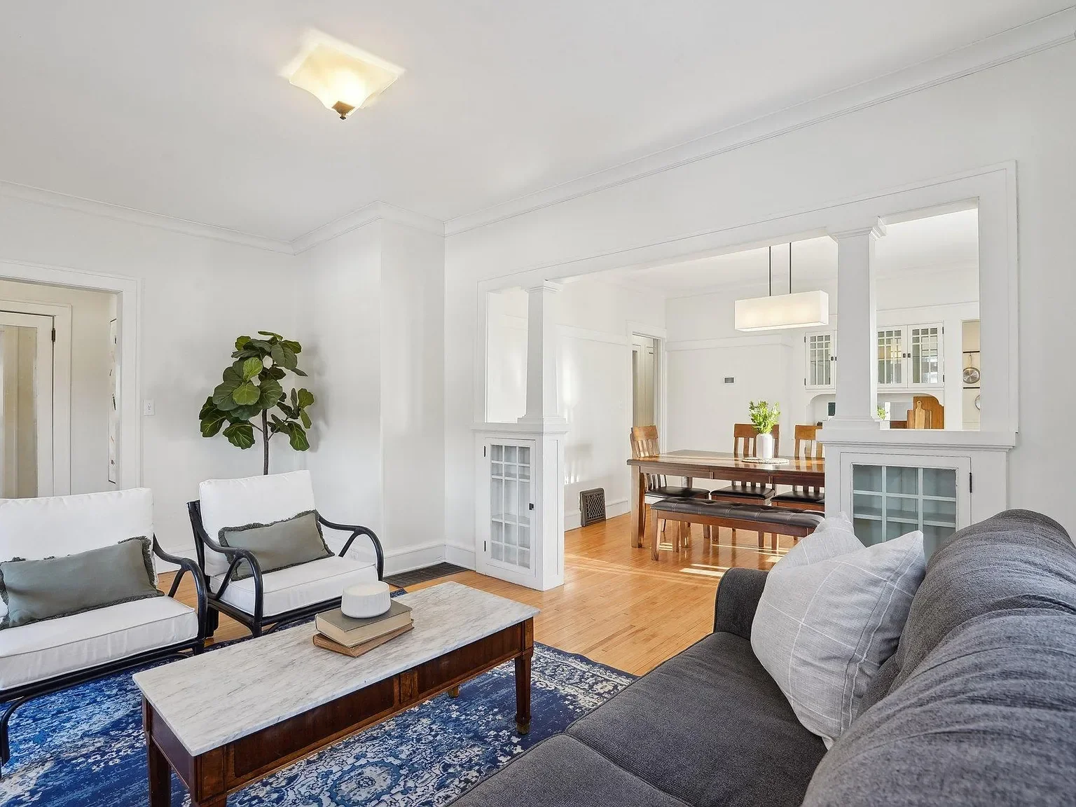 Living room with smoky gray sofa, white and black armchairs, a coffee table, a blue patterned rug, and a tall green plant, with a dining area visible through a wide opening.