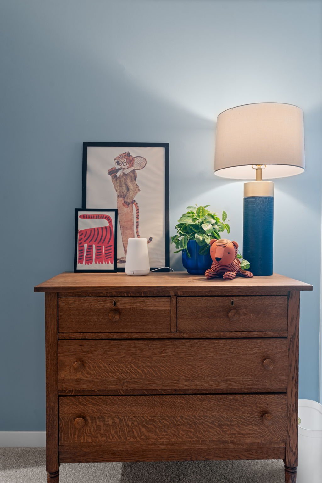 A wooden dresser with framed art, a small white diffuser, a green plant in a blue pot, a plush tiger toy, and a large table lamp with a beige shade against a light blue wall.