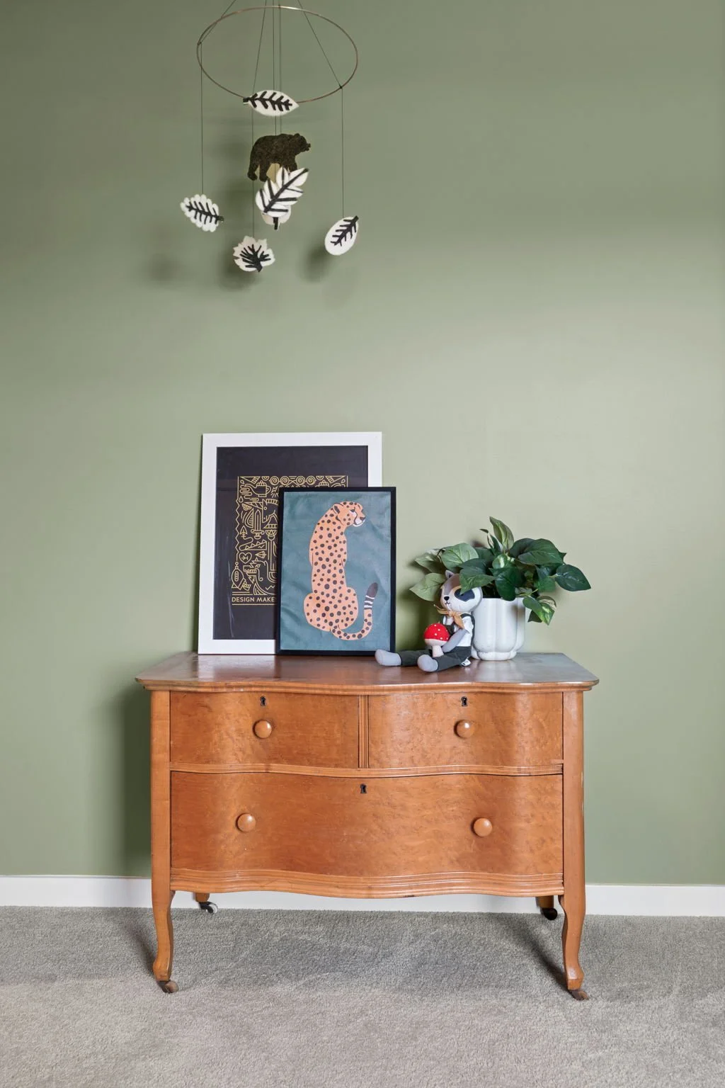 A wooden dresser against a light green wall with framed artwork, a potted plant, and a plush toy on top. A mobile with black and white leaves and a bear hangs above.