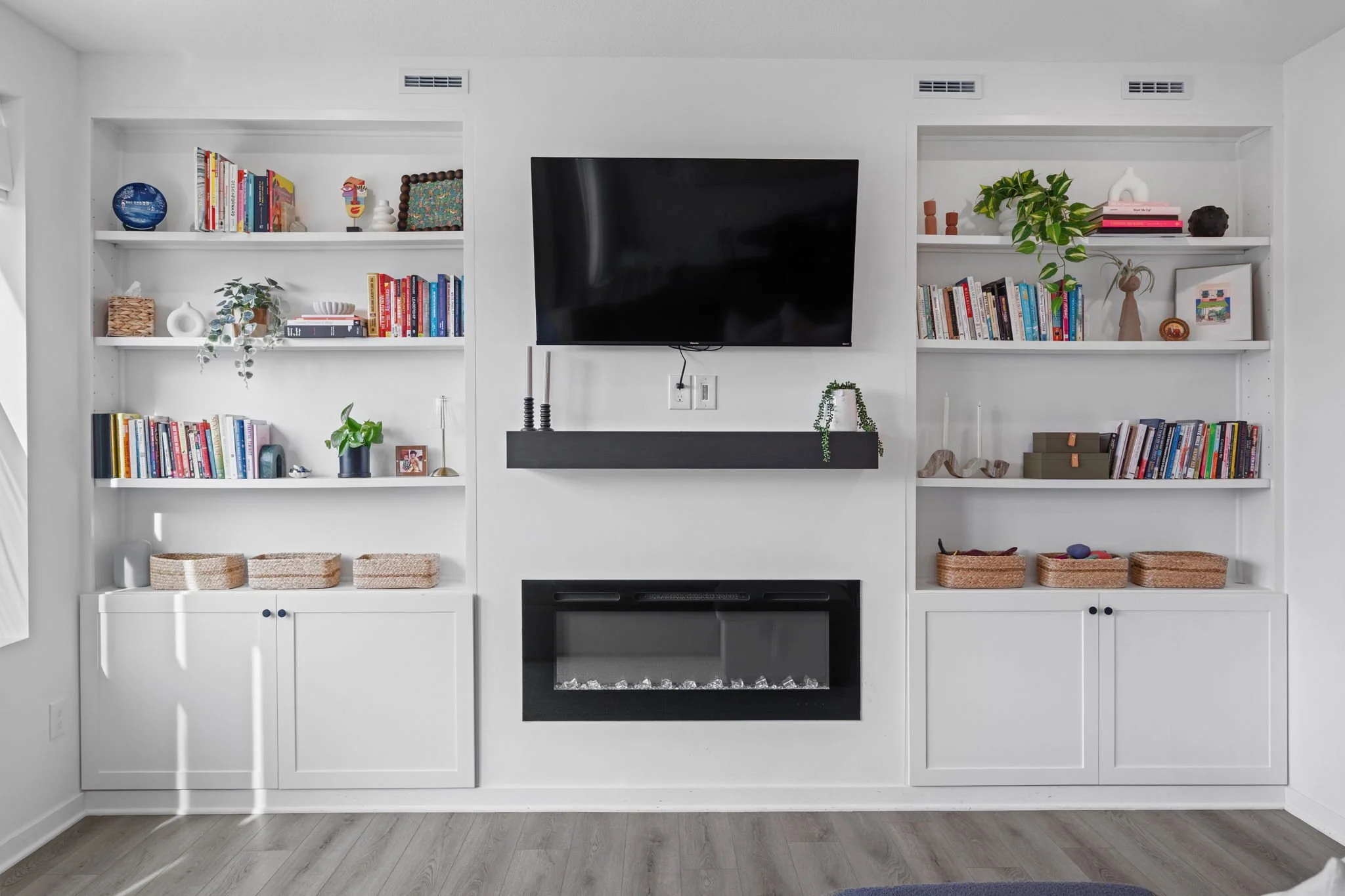 White built-in shelves with books, decorative items, and plants flanking a wall-mounted TV above a modern fireplace in a living room.