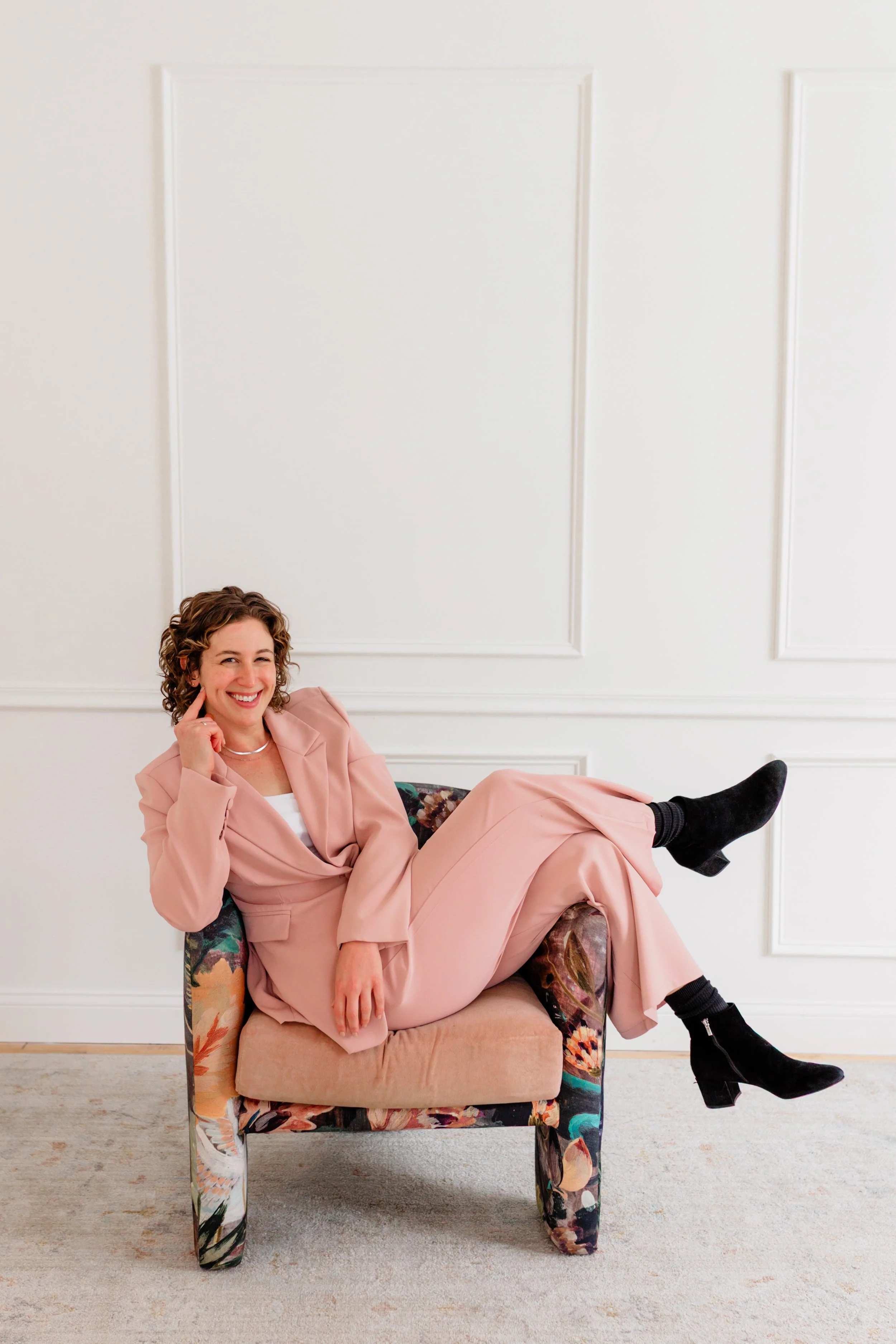 A woman in a pink suit sitting on a colorful armchair, smiling and posing with her legs crossed in a bright, minimalist room.