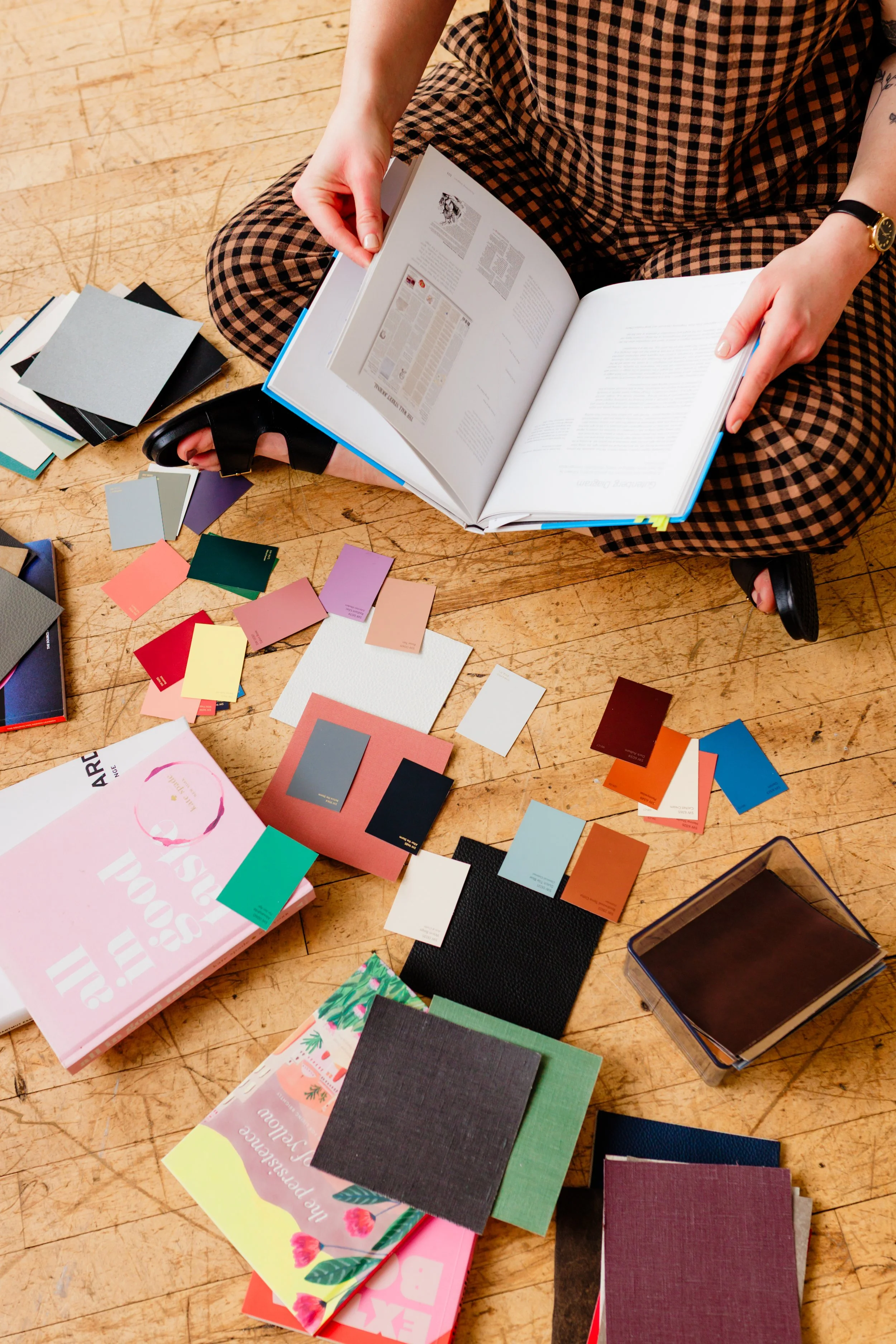 Person sitting cross-legged on a wooden floor, looking at an open book, surrounded by various colored sample cards, booklets, and fabric swatches.