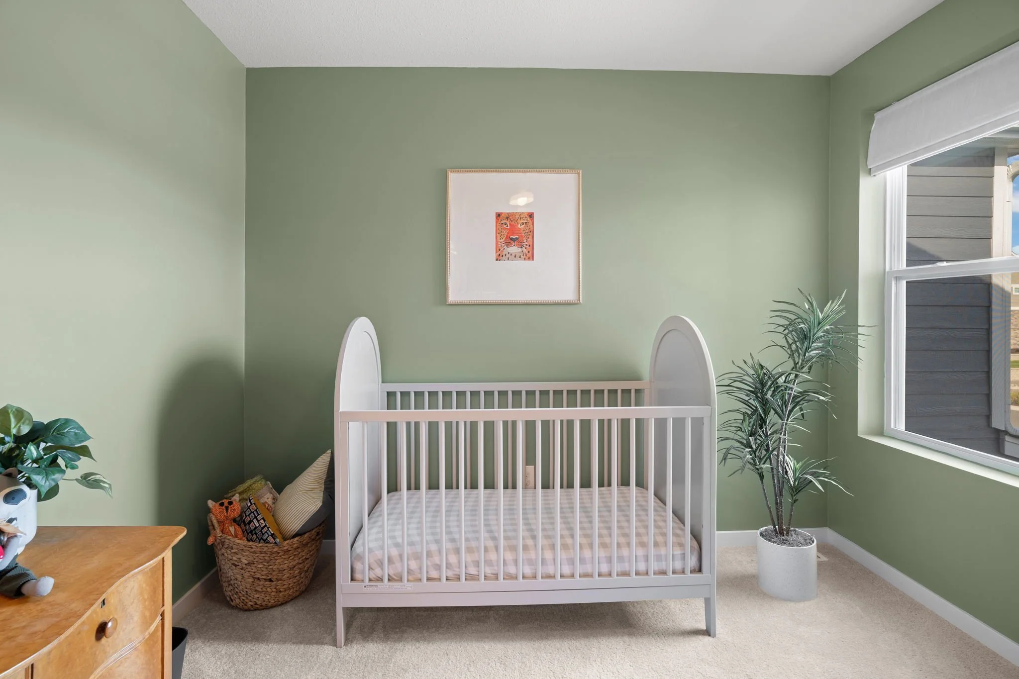 A nursery with green walls, a white crib, a window with a view outside, a plant in a white pot, and wall art of a lion.