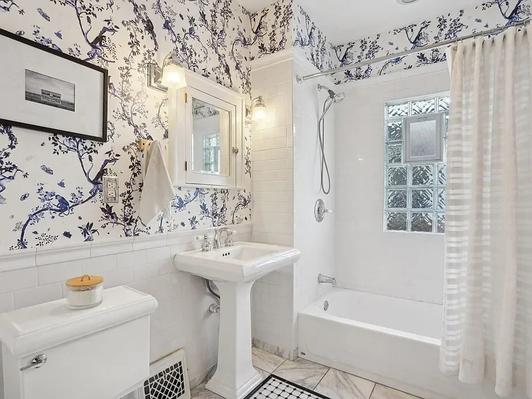 Bathroom with white tile walls, blue and white patterned wallpaper above the tiles, a white pedestal sink, a wall-mounted mirror, a framed black and white photo, a white toilet with a jar on top, a small window with frosted glass blocks, and a bathtu