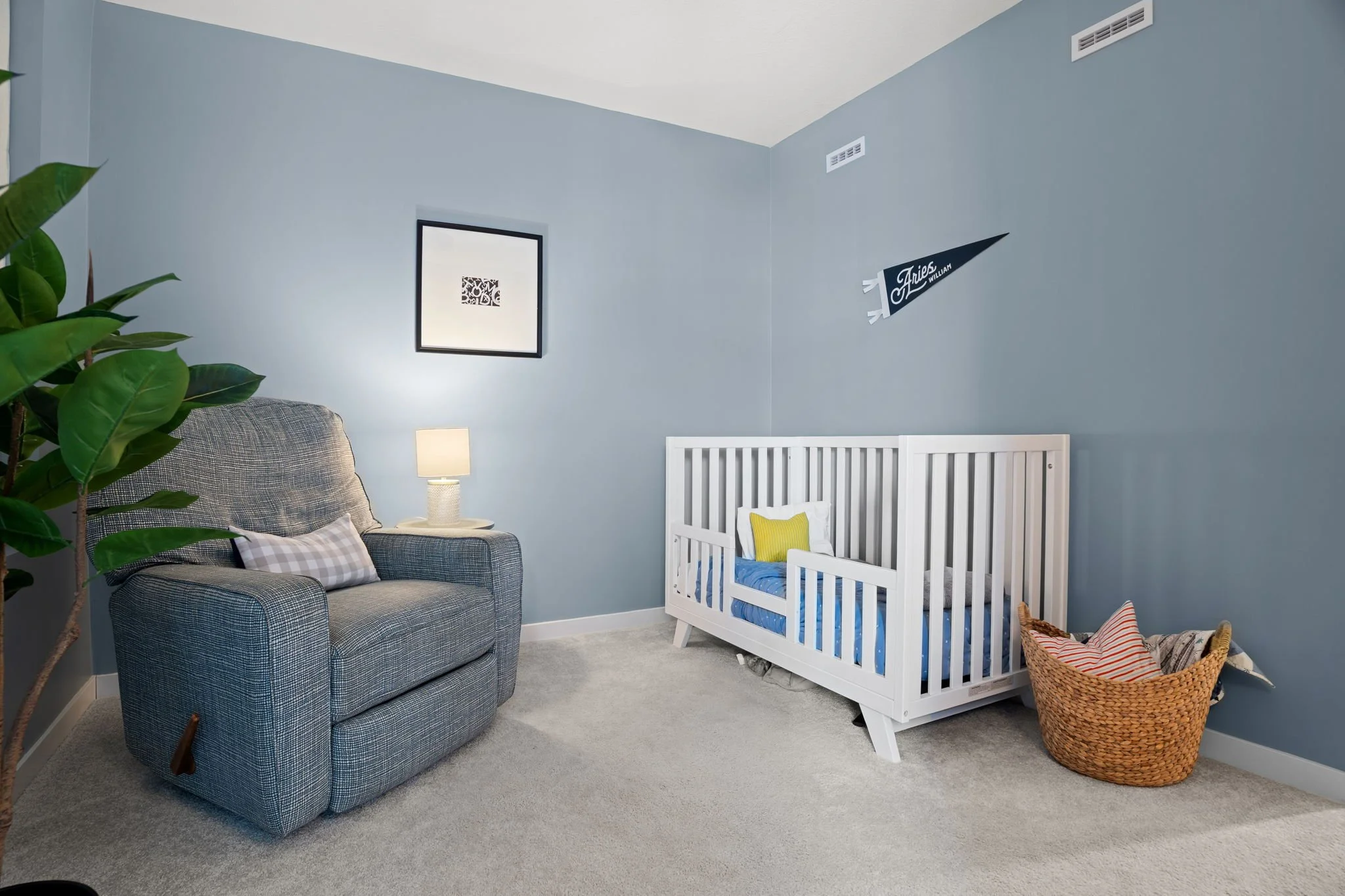 Living room with gray armchair, white crib, wicker basket, blue walls, framed art, plant, and a lamp.