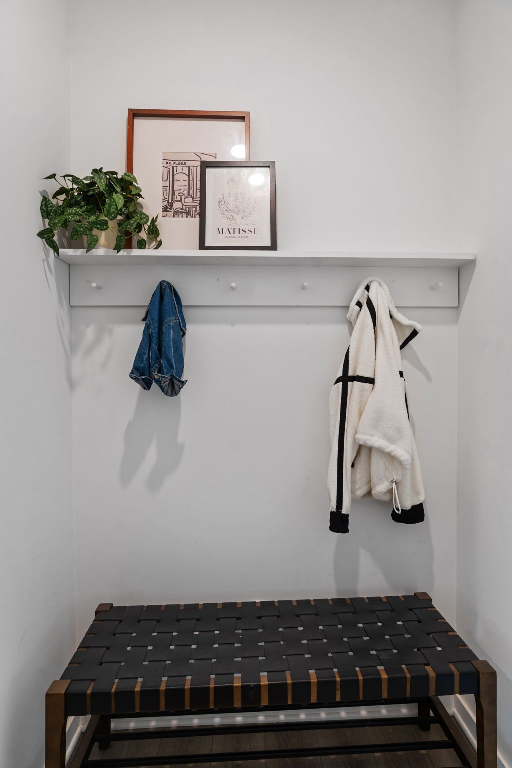A minimalist entryway with a white wall, a narrow white shelf with hooks, a potted plant, framed artwork, and a bench with a woven black and brown seat.