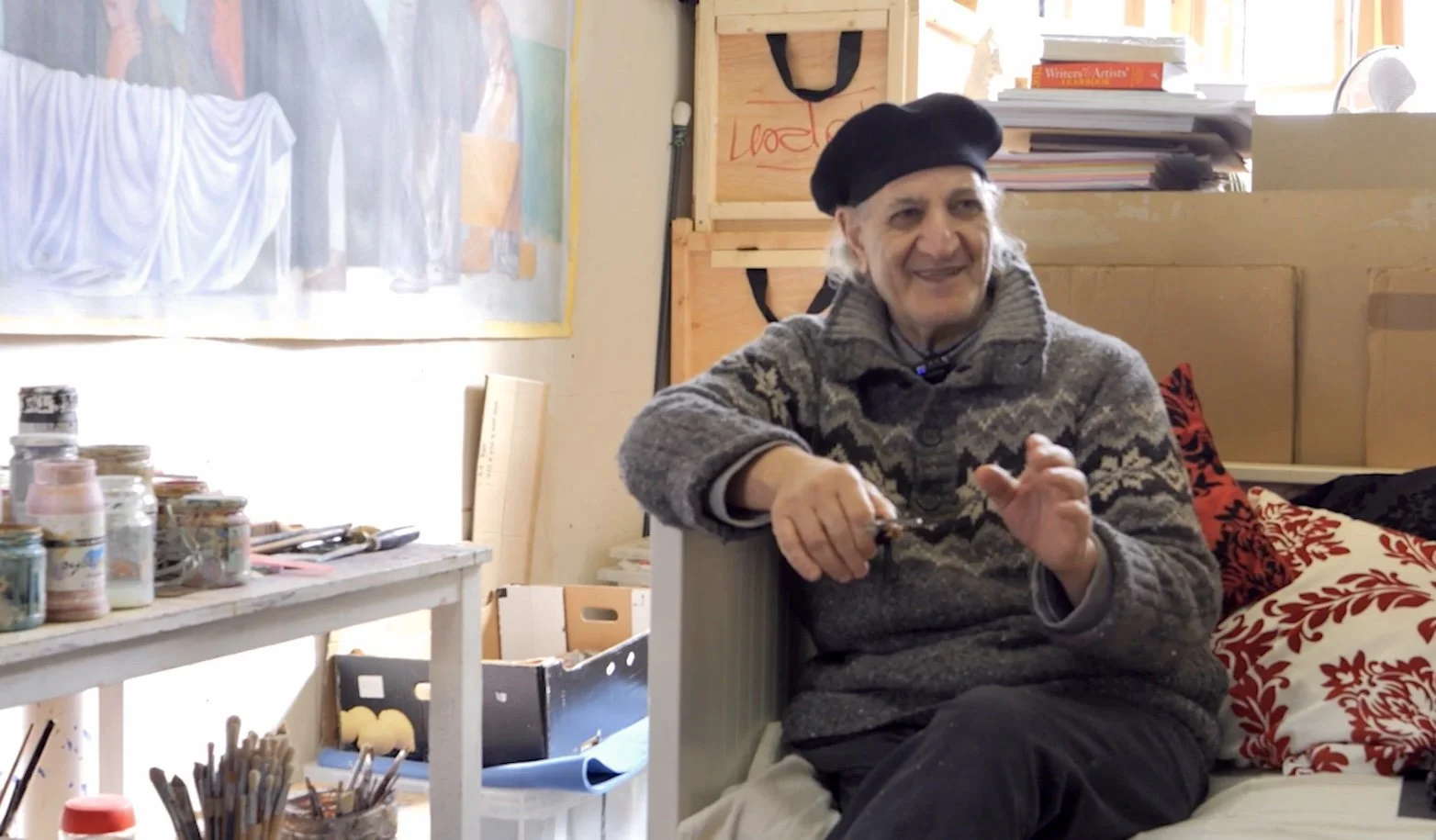 An older man sitting on a bed with colorful throw pillows, wearing a patterned sweater and a black beret, smiling and possibly holding a pen or similar object, in a cozy, art-filled room with shelves, books, and art supplies.