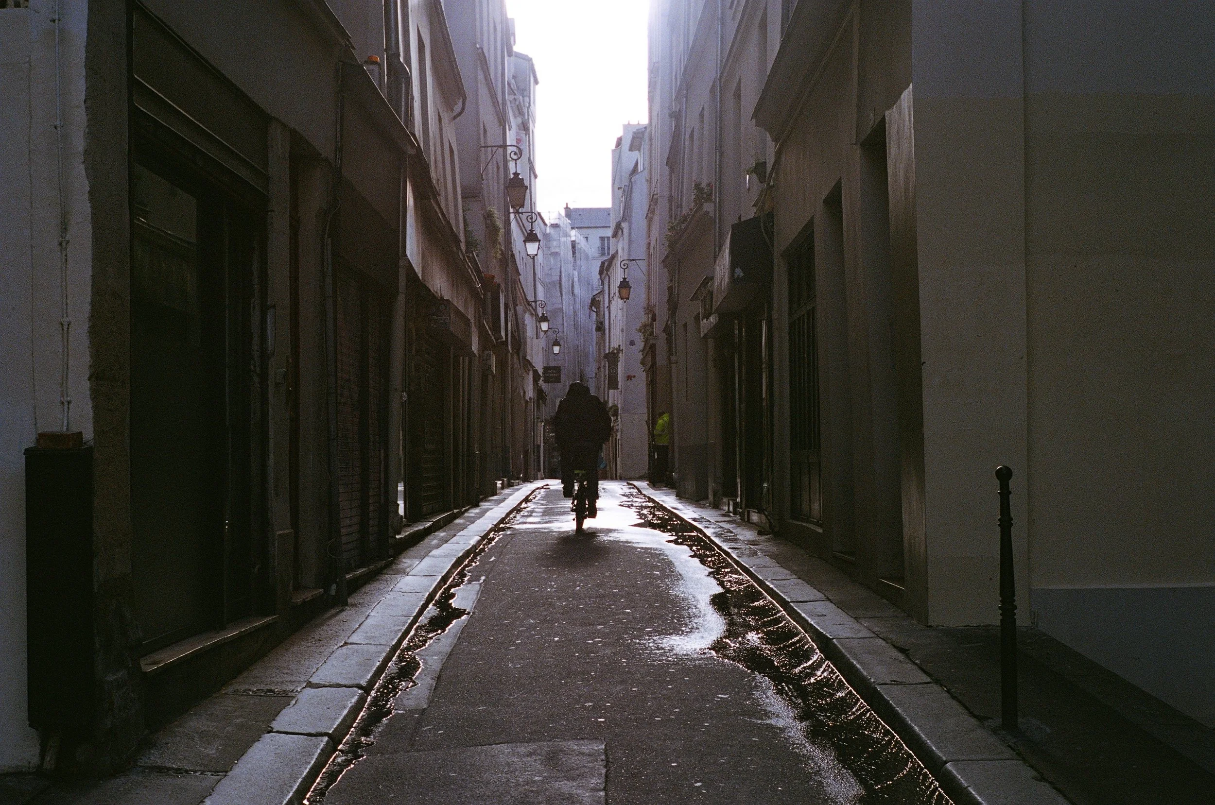 A narrow alleyway in a city with tall buildings on both sides, sunlight shining at the end, a person riding a bicycle away from the camera in the center, and a person in a yellow jacket further down the alley.