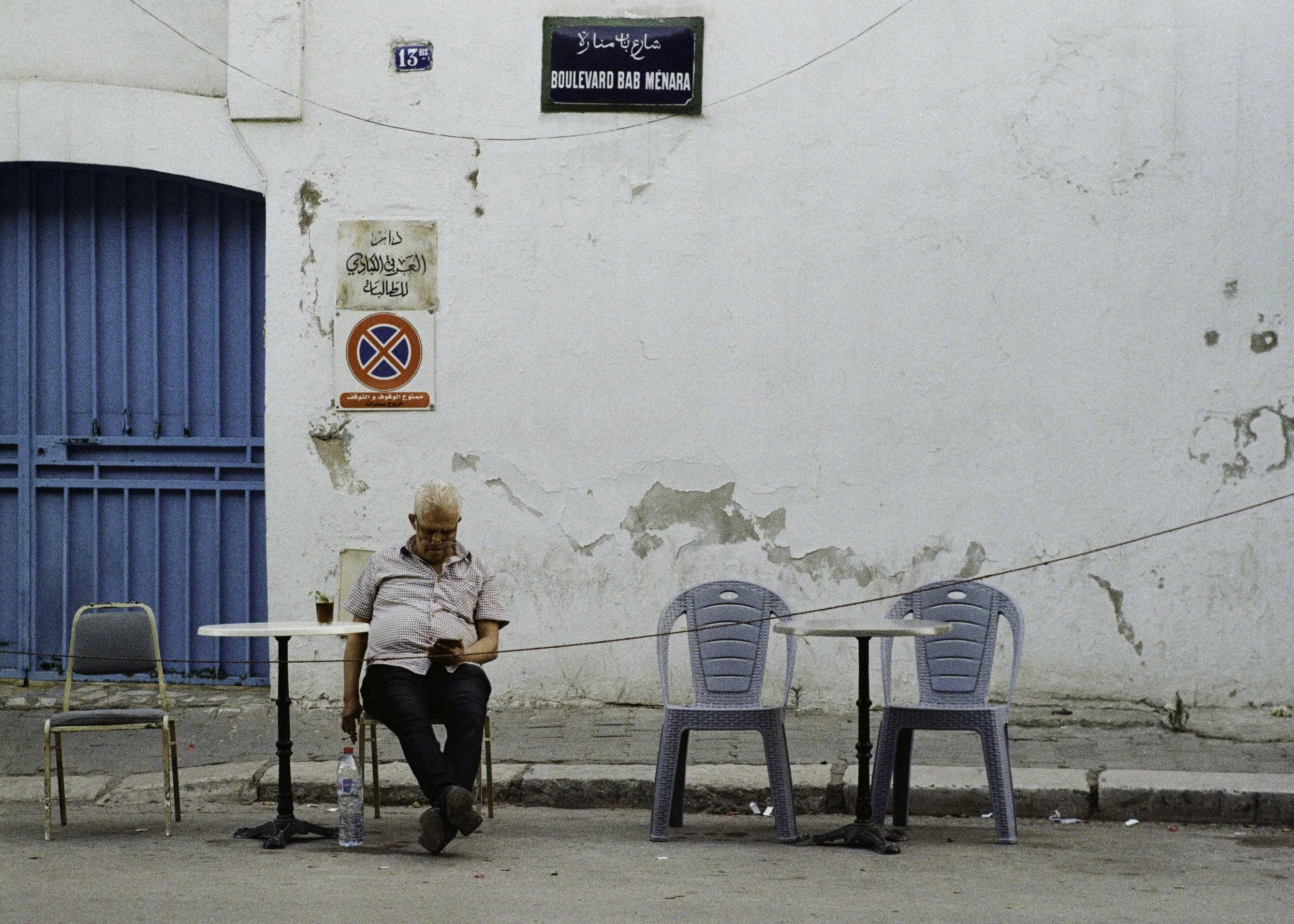 An elderly man with glasses, wearing a plaid shirt and dark pants, sitting on a chair on the sidewalk, looking at his phone. There are three empty chairs and two small tables; one table has a small pot with a plant. The wall behind is white with peel