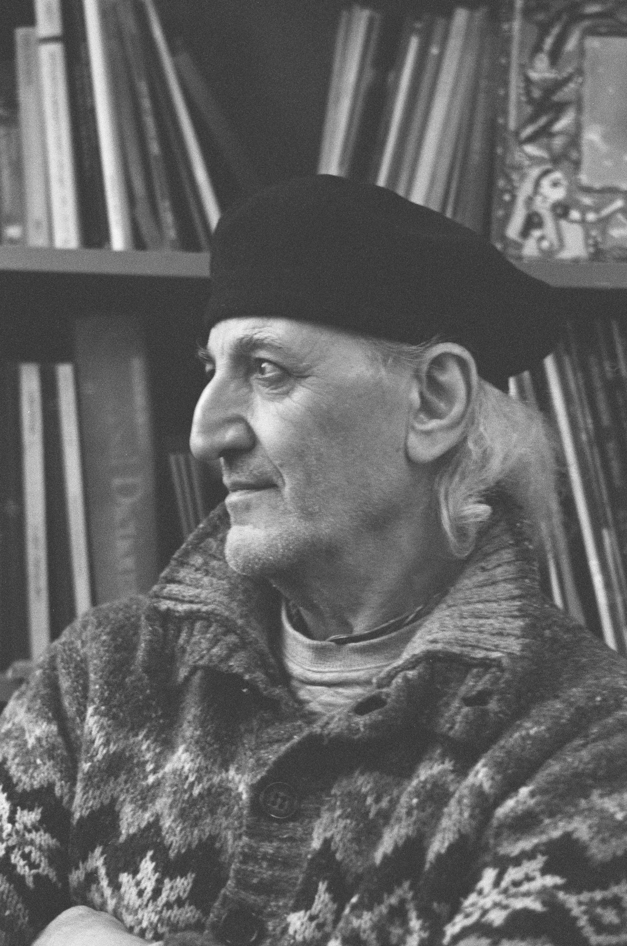Black and white photo of an elderly man with long, wavy hair, wearing a patterned sweater and a beret, sitting in front of a bookshelf filled with books.