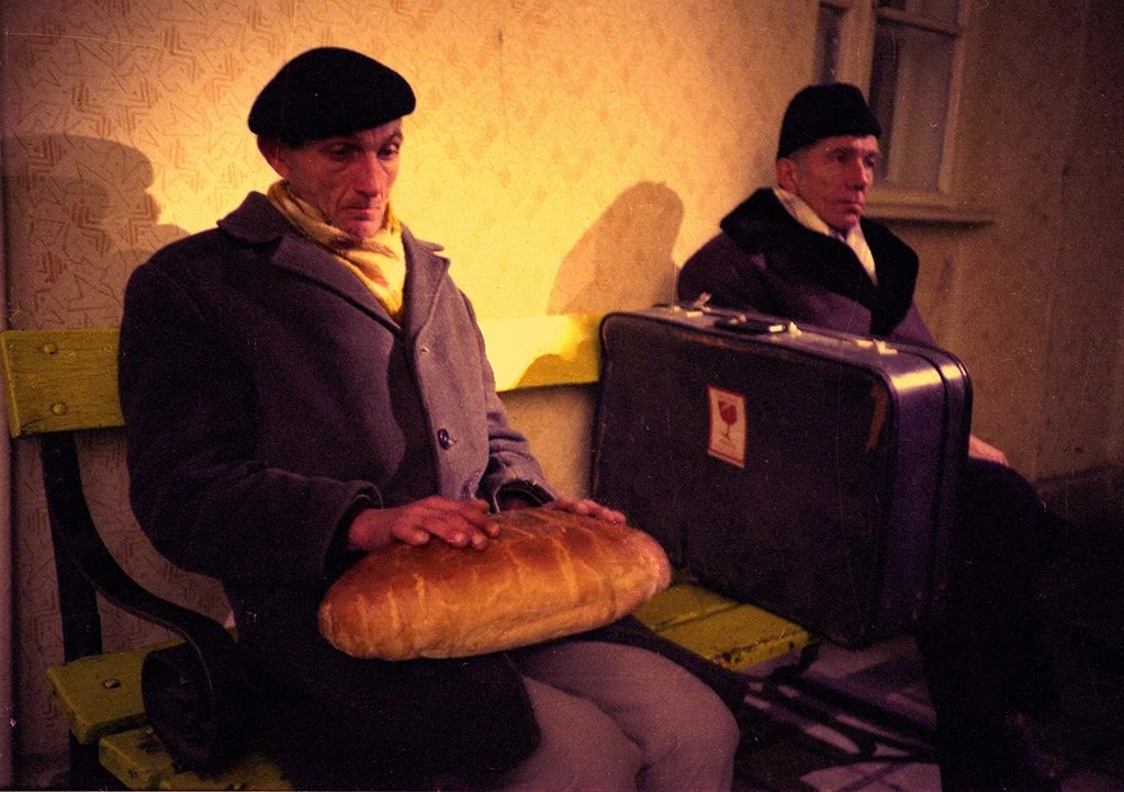 Two men sitting on a yellow bench in a room with patterned wallpaper, one holding a loaf of bread and the other with a suitcase.