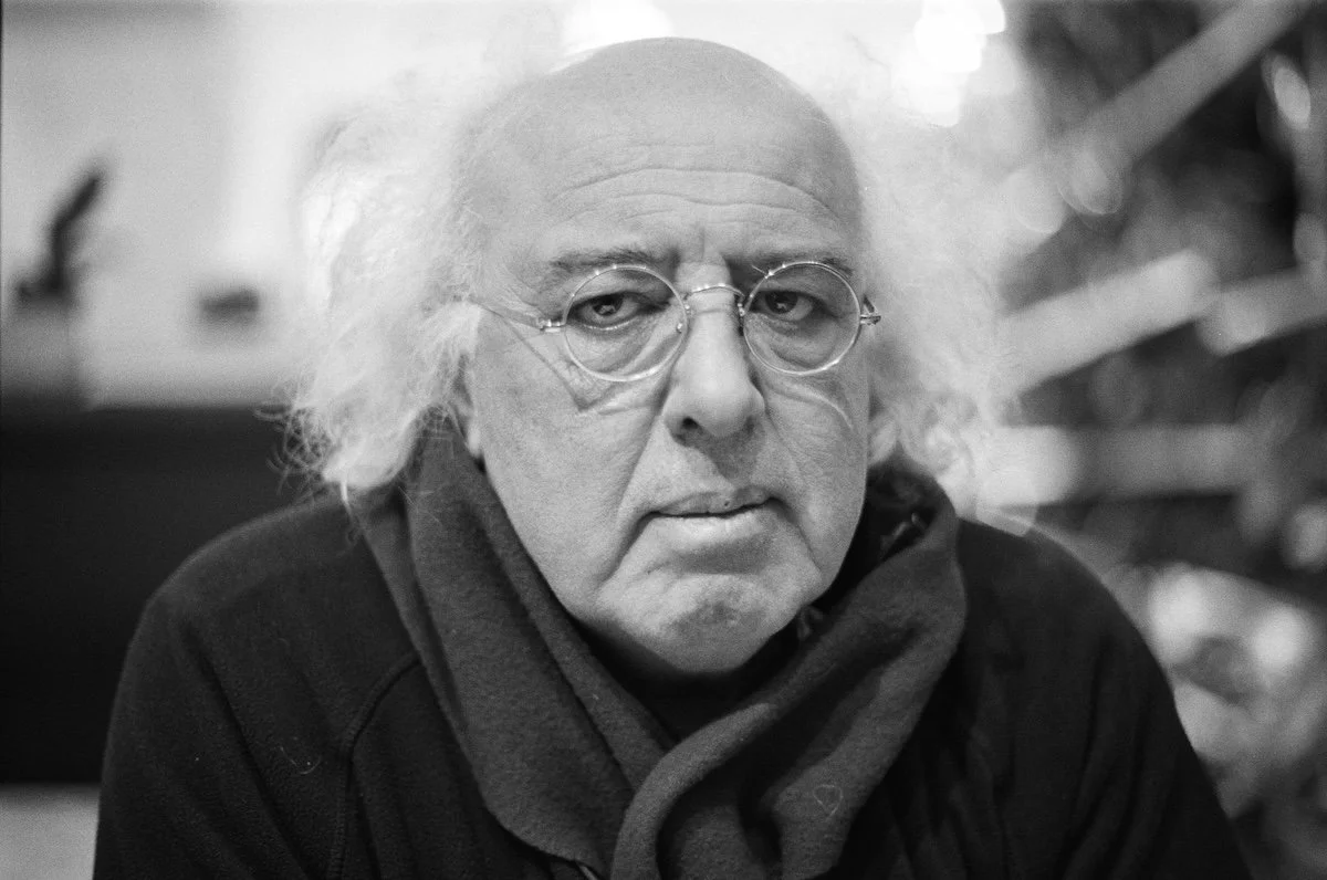 A black and white photo of an older man with long, disheveled white hair, round glasses, and a serious expression. He is wearing a dark hoodie and a scarf, with a blurred background.