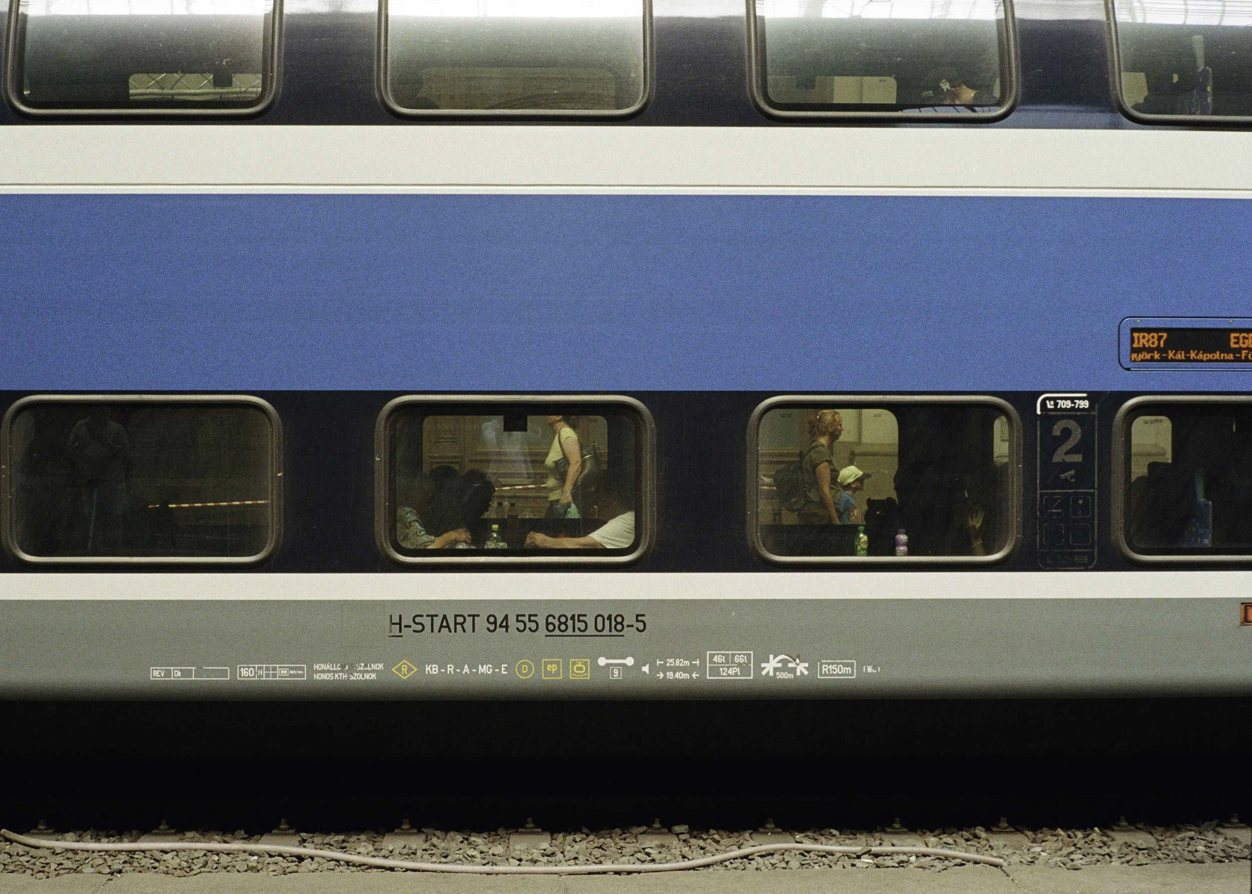 Side view of a modern double-decker train with passengers visible through its windows, some sitting and some standing, with a blue and gray exterior, rail track below, and a digital sign indicating train route to Eger.