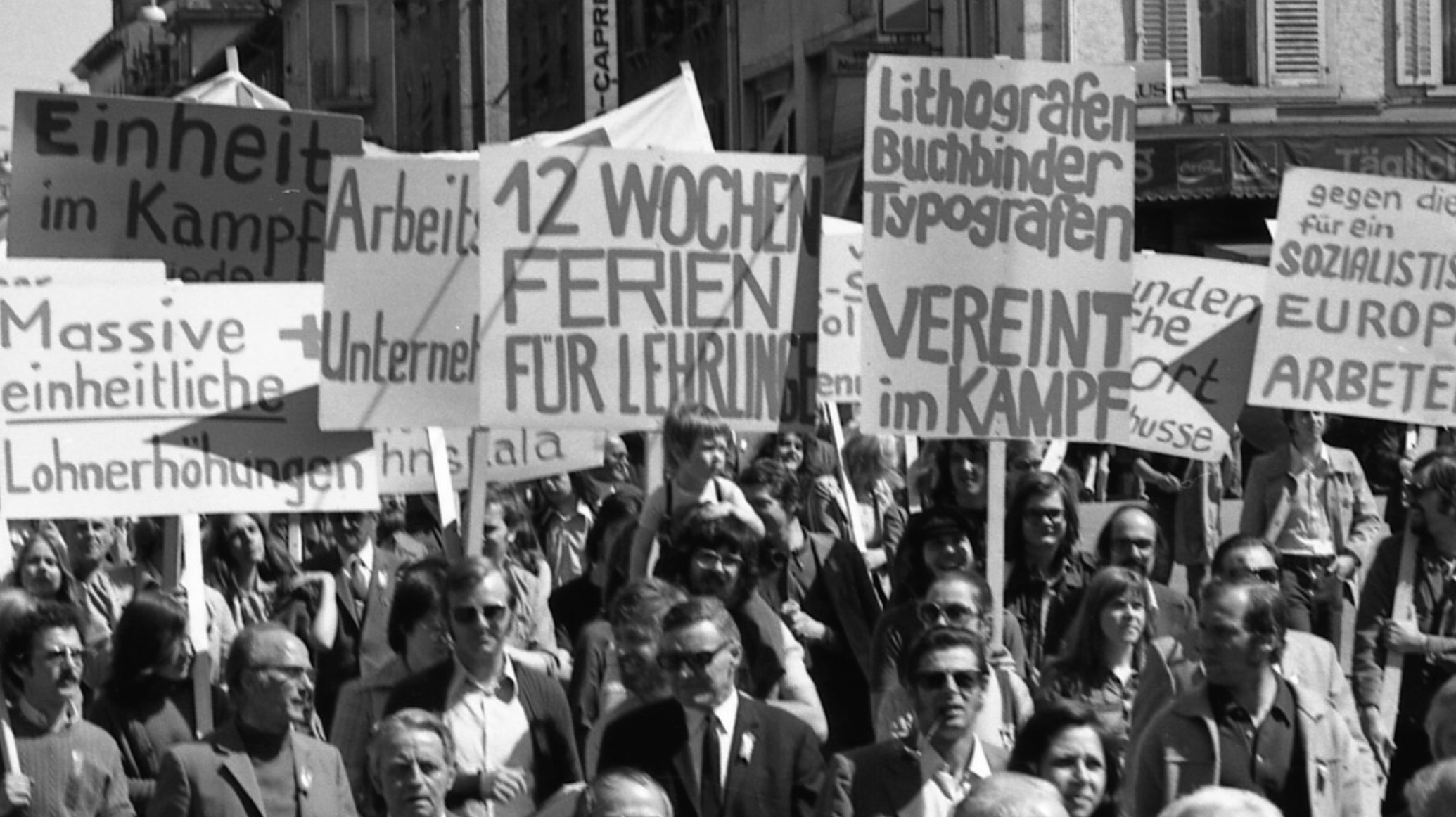 A black and white photo of a crowd of people at a protest holding signs and banners with German text, advocating for workers' rights, education, and social issues.
