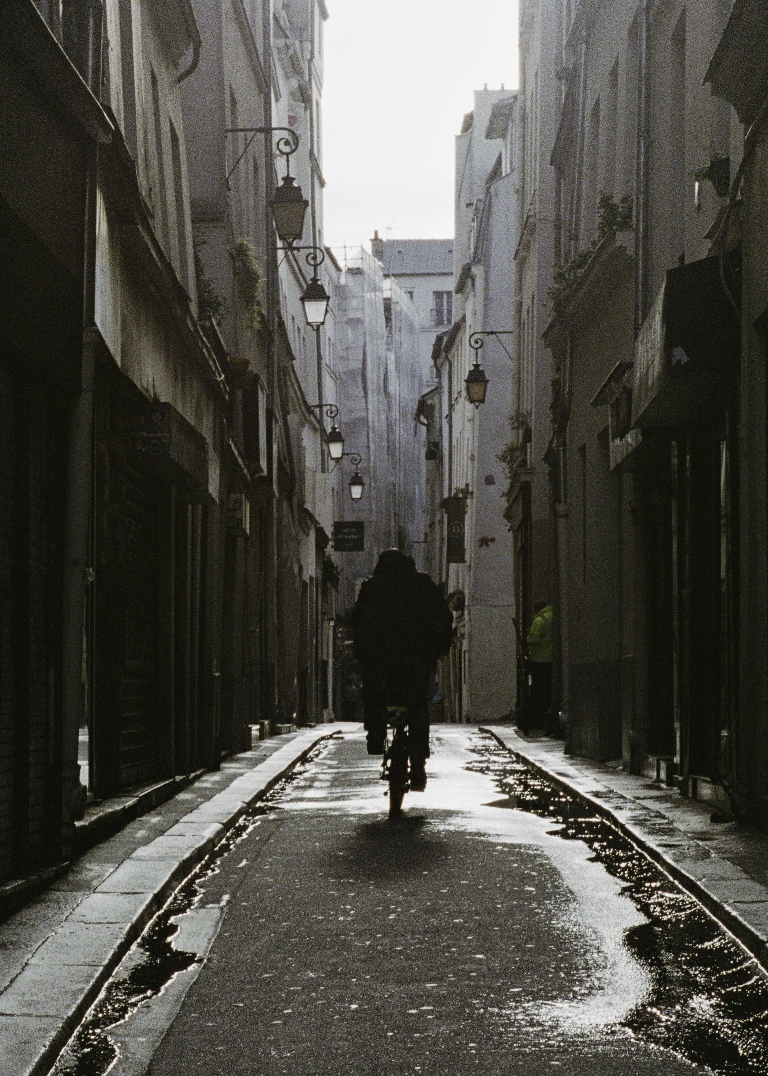 A person riding a bicycle down a narrow Parisian alley illuminated by street lamps, with buildings on either side and a bright sky above.