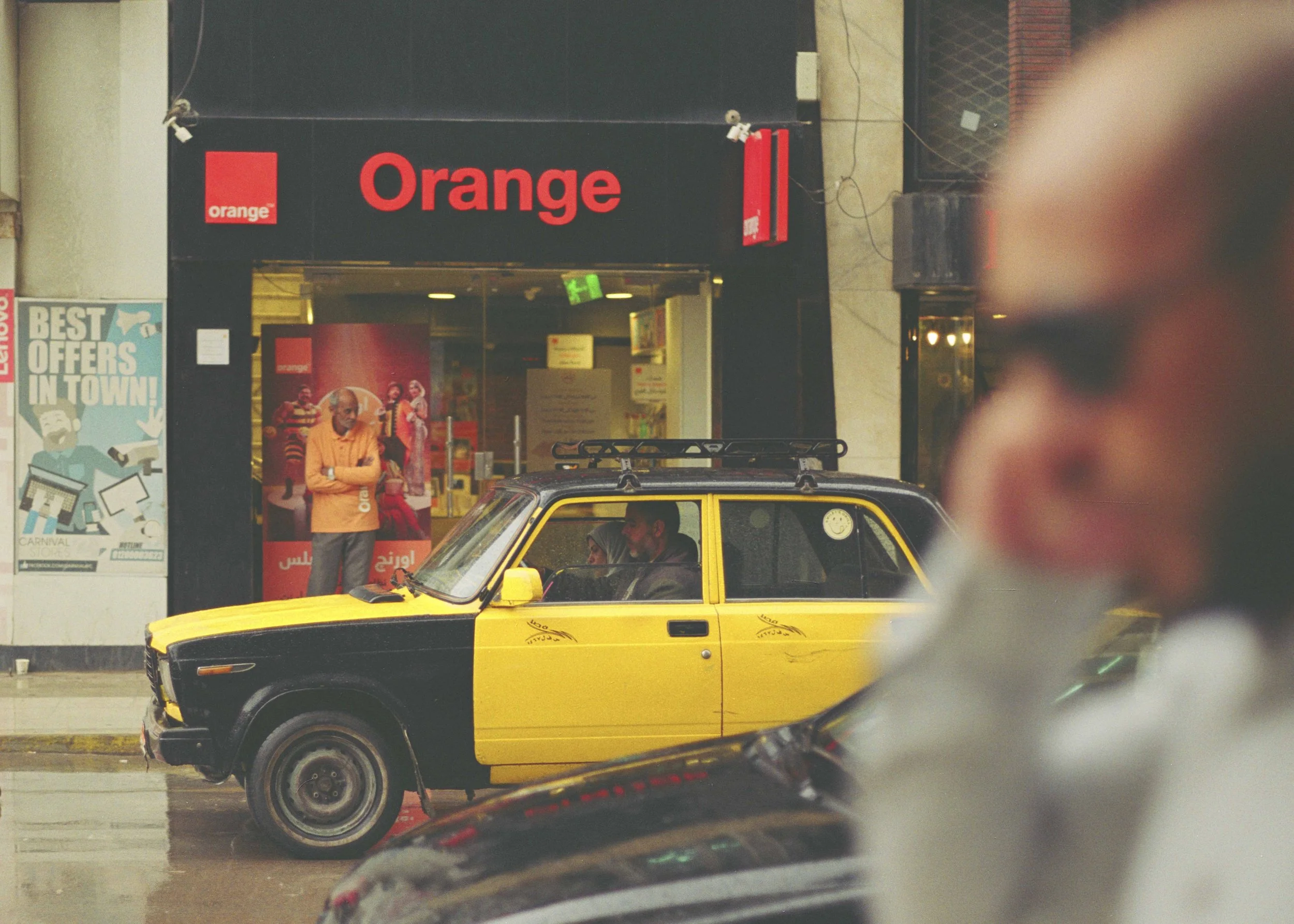 A yellow and black car parked on a street in front of an Orange store. There are two people sitting inside the car, and a man standing outside the store looking inside. The store has a black and red sign with the logo and name 'Orange'. A person in t