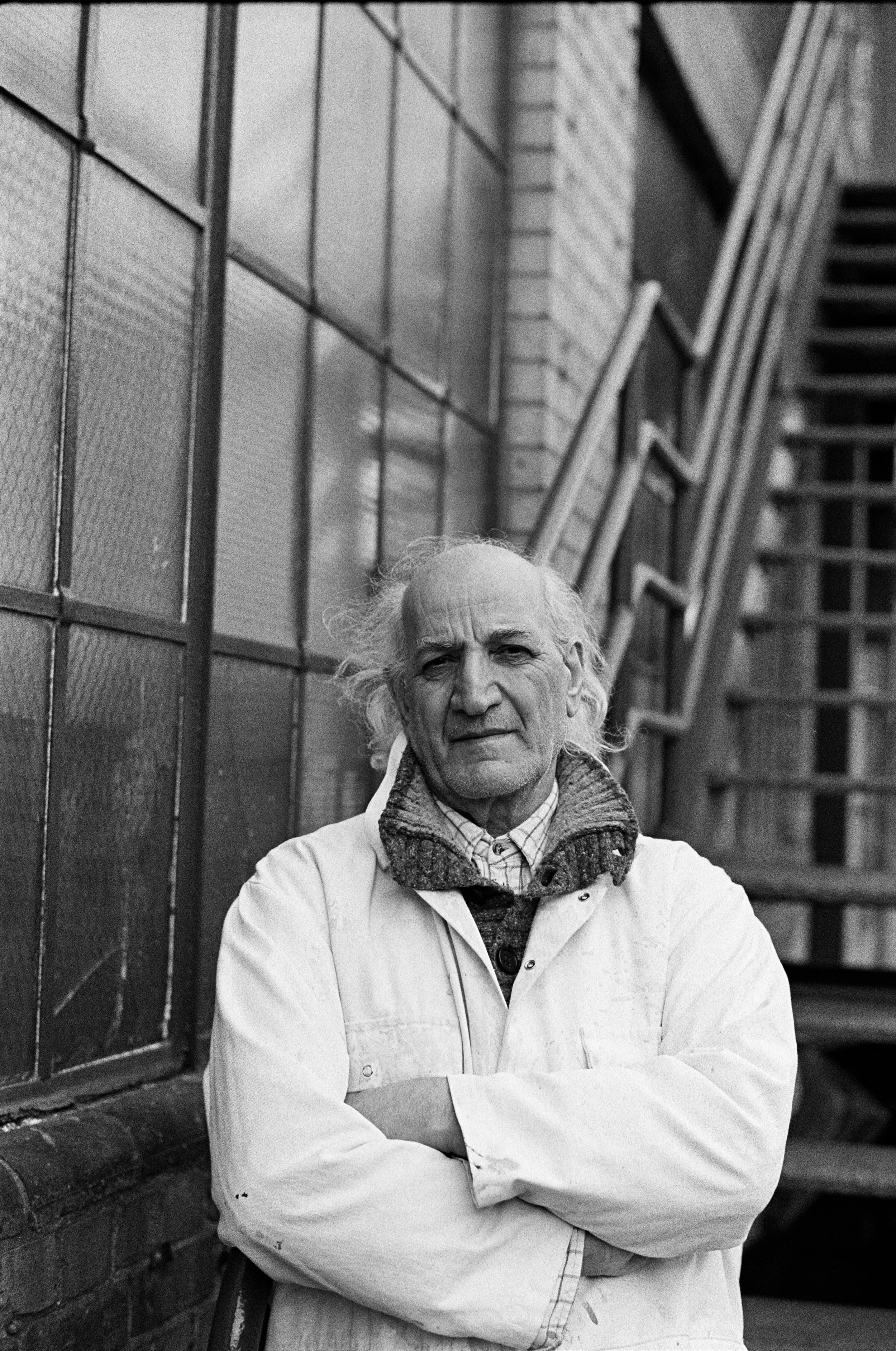 An elderly man with white hair and a serious expression, wearing a white coat and a sweater, standing outdoors near a brick and glass building with stairs.