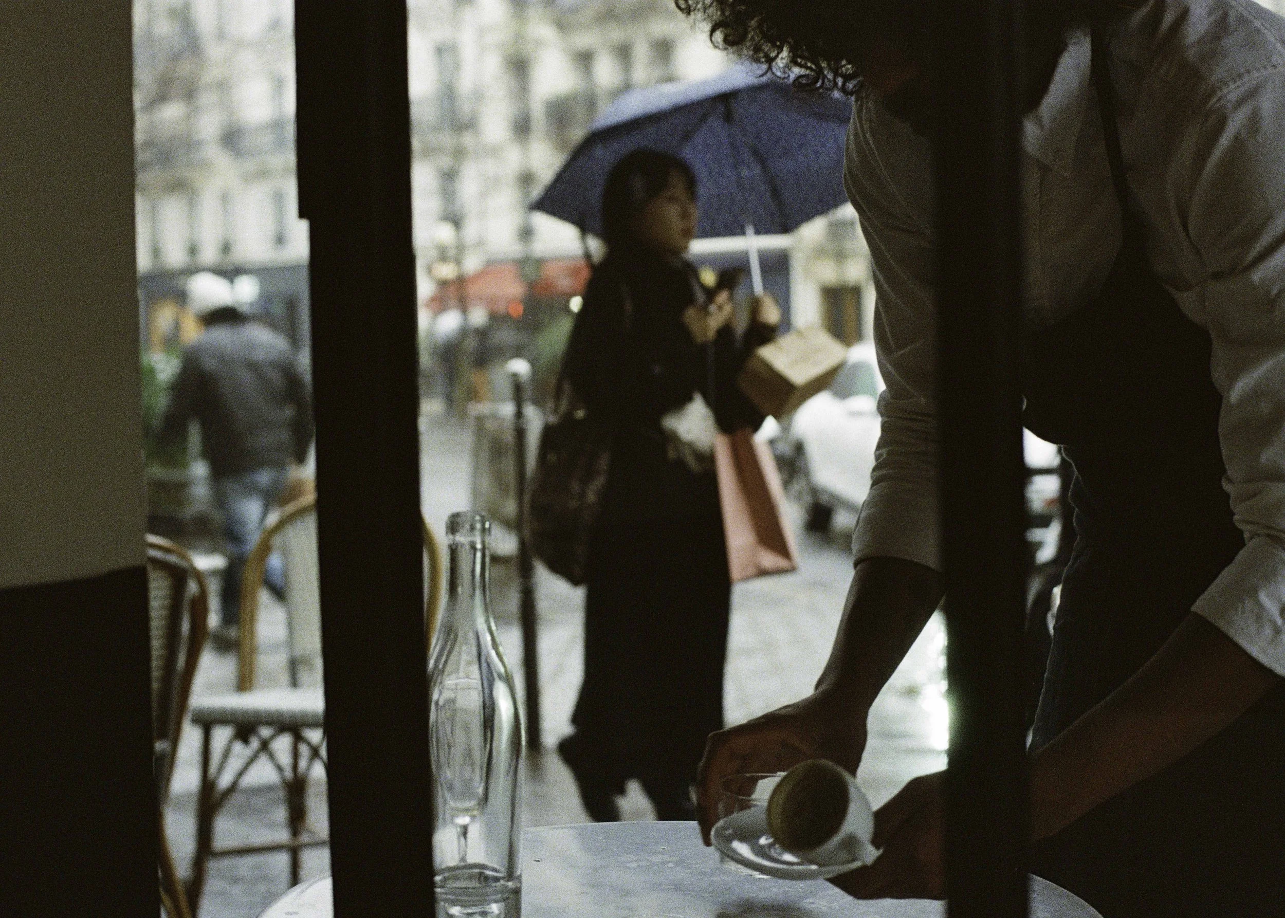 Person pouring a drink at a table inside a cafe while people with umbrellas walk outside on a rainy day.