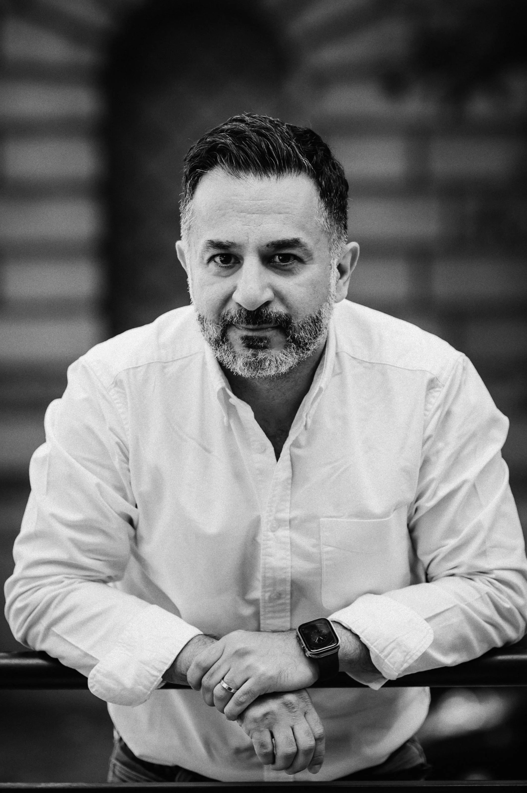 Black and white portrait of a man with short dark hair and a beard, wearing a white button-up shirt and a smartwatch, leaning on a railing, looking directly at the camera.