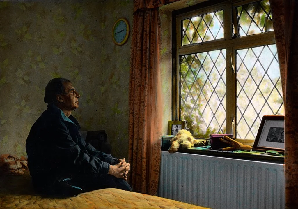 An elderly woman sitting on a bed in a dimly lit room, looking out the window. The window has a diamond-shaped grid pattern with curtains on both sides. There are framed photographs, a teddy bear, and a pair of yellow gloves on the windowsill.