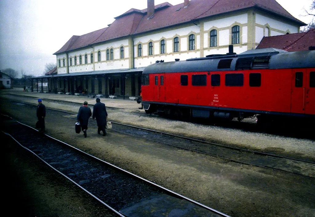 Three men walking on a train station platform with a red train and a large yellow building in the background.