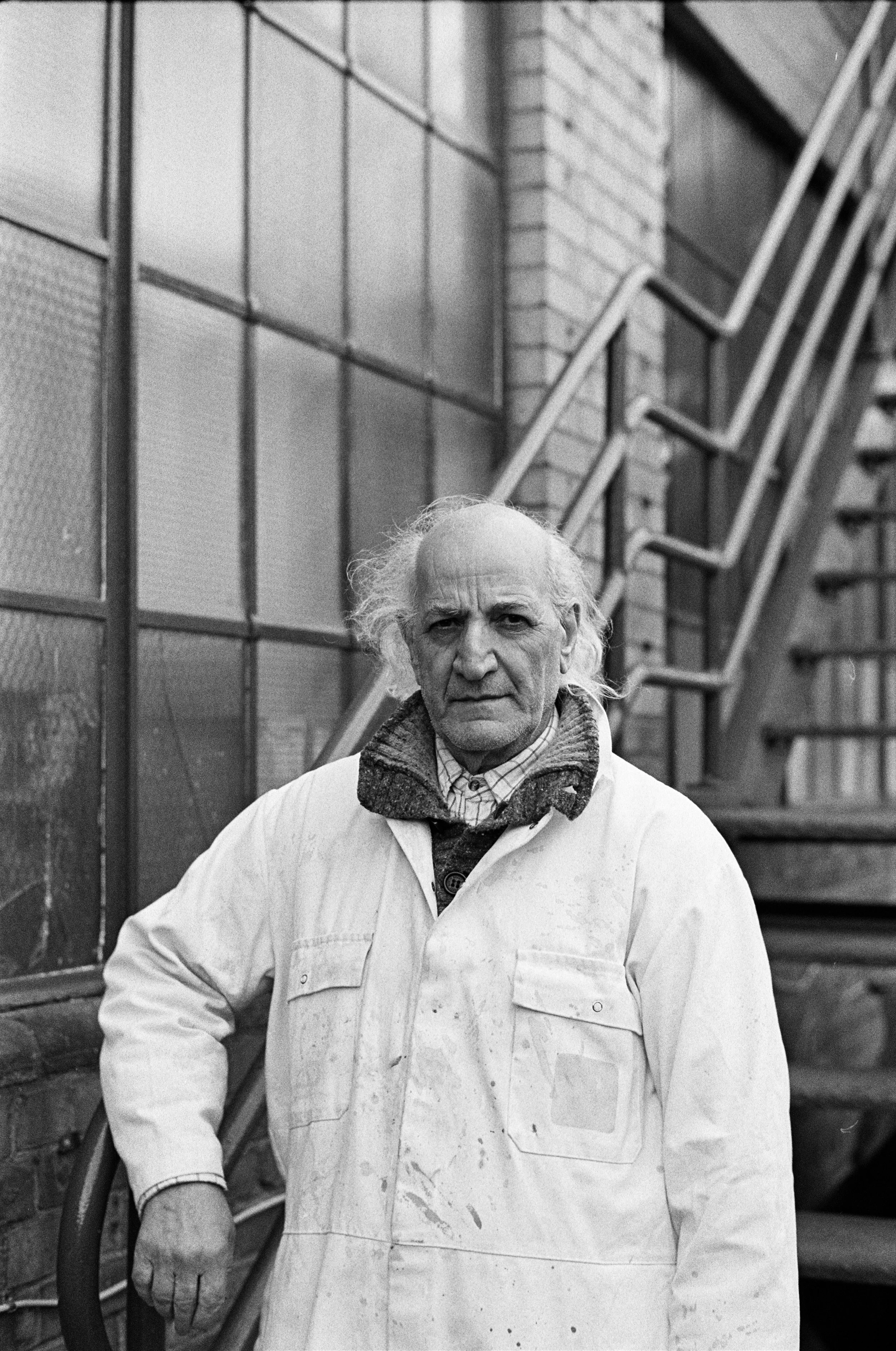 A black-and-white photo of an elderly man with long, frizzy hair and a serious expression, wearing a white lab coat with stains and a scarf, standing outdoors near a brick building with large windows and a staircase.