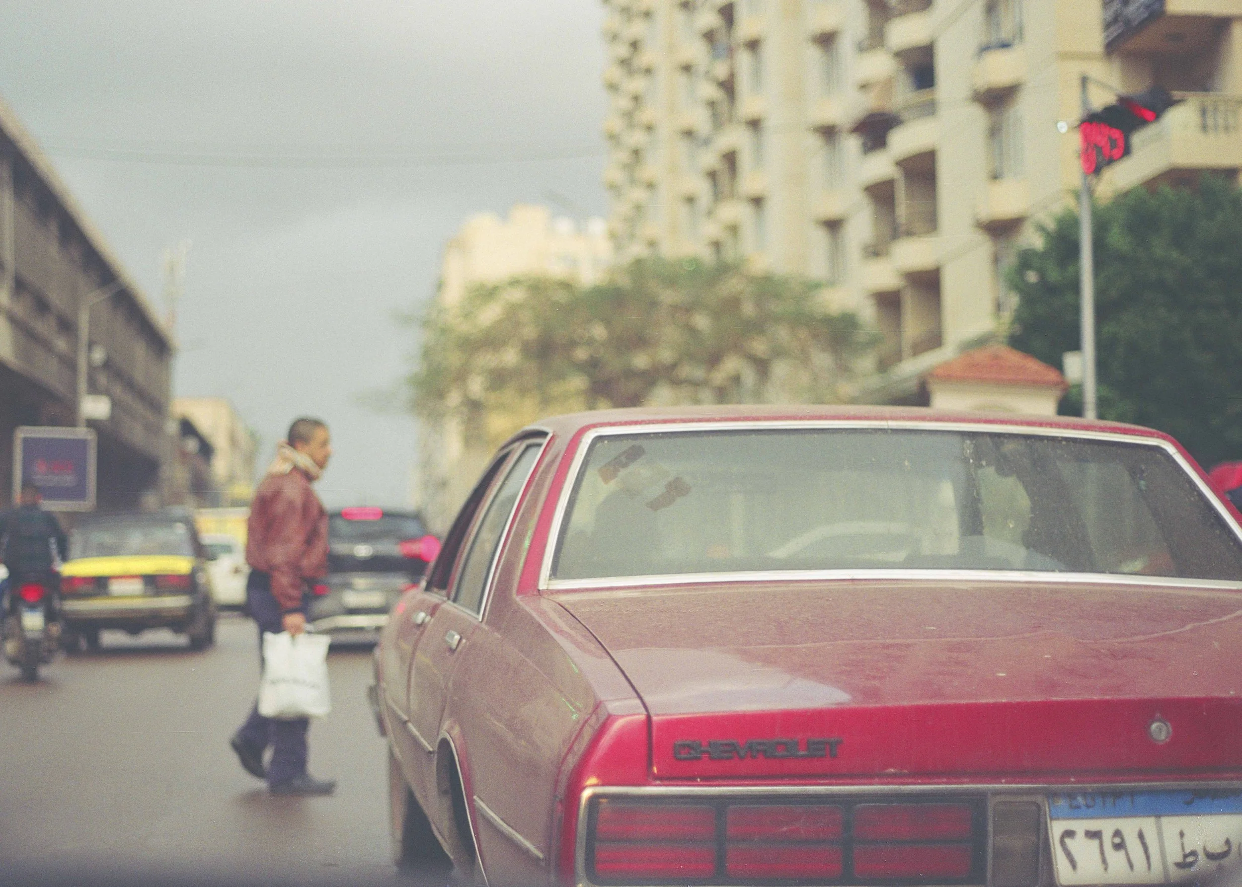 A man crossing a street with cars parked along the side, a red Chevrolet car in the foreground, buildings in the background, and a traffic light showing red.